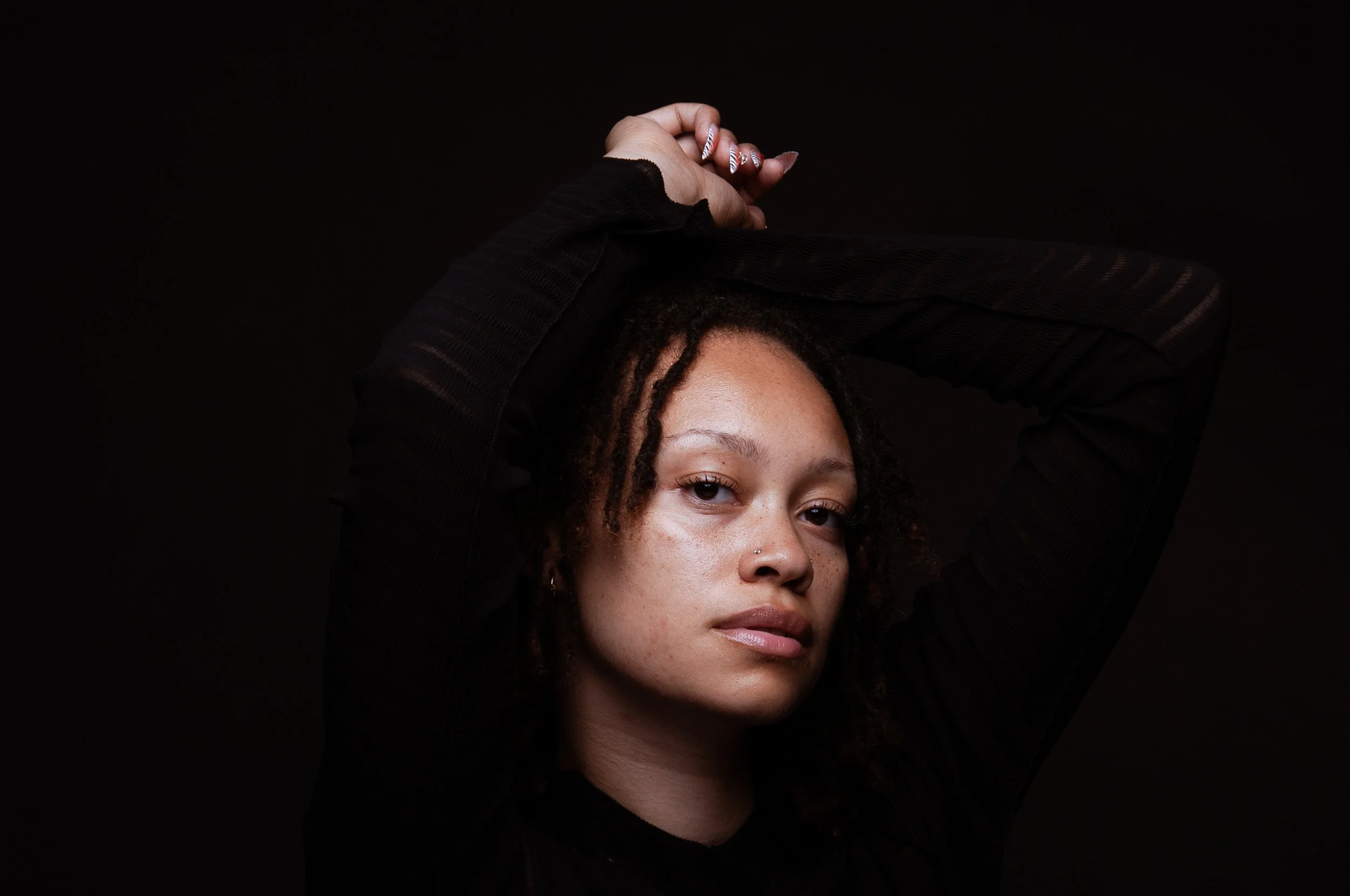 Close-up portrait of a woman with curly hair, nose piercing, and patterned nails posing in black clothing against a dark background