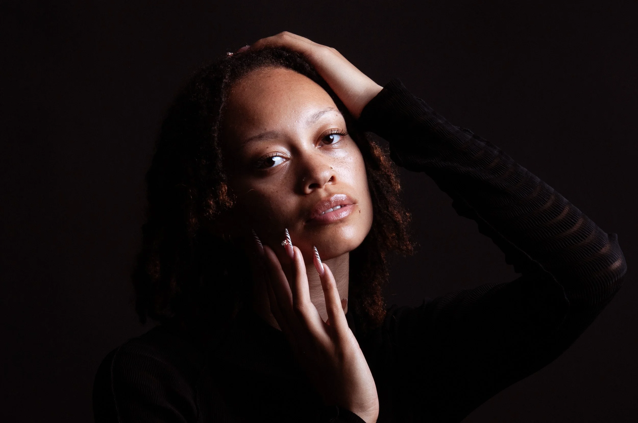 Close-up portrait of a Black woman with curly hair, long manicured nails, and a black outfit posing against a dark background