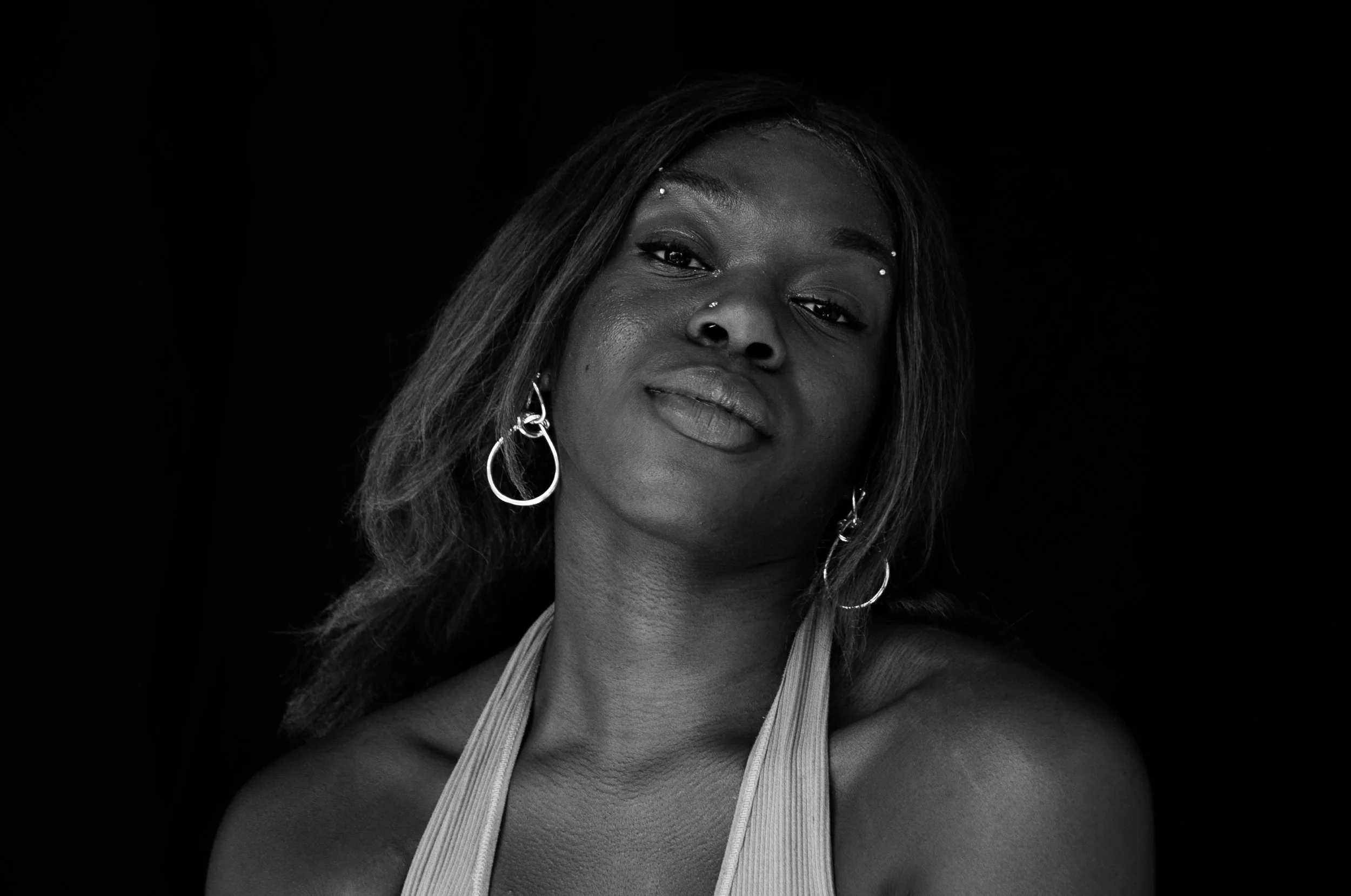 Black-and-white close-up portrait of a woman with hoop earrings, facial jewelry, and long hair against a black background