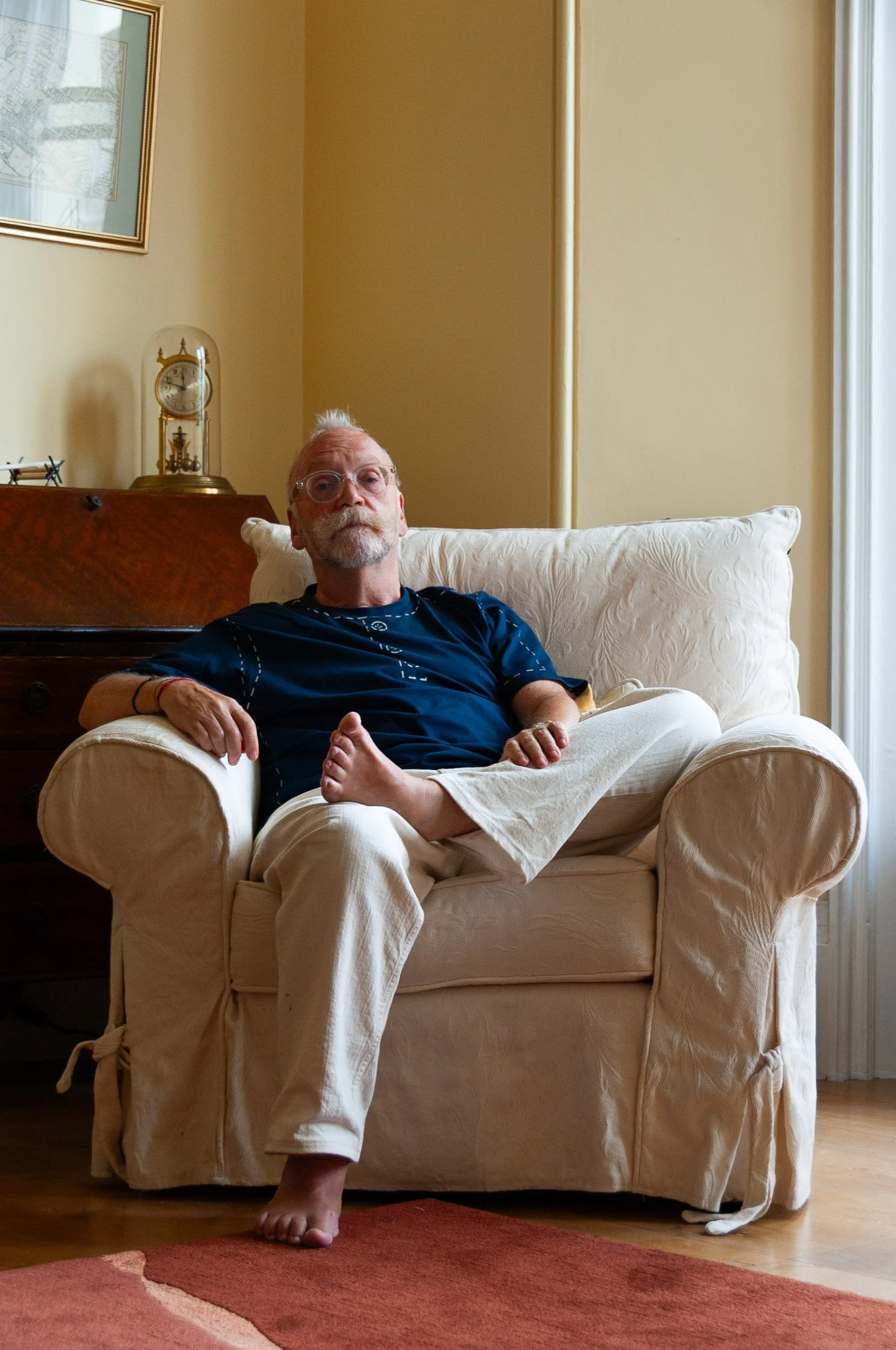 Older man wearing glasses sits barefoot in a white armchair at home.