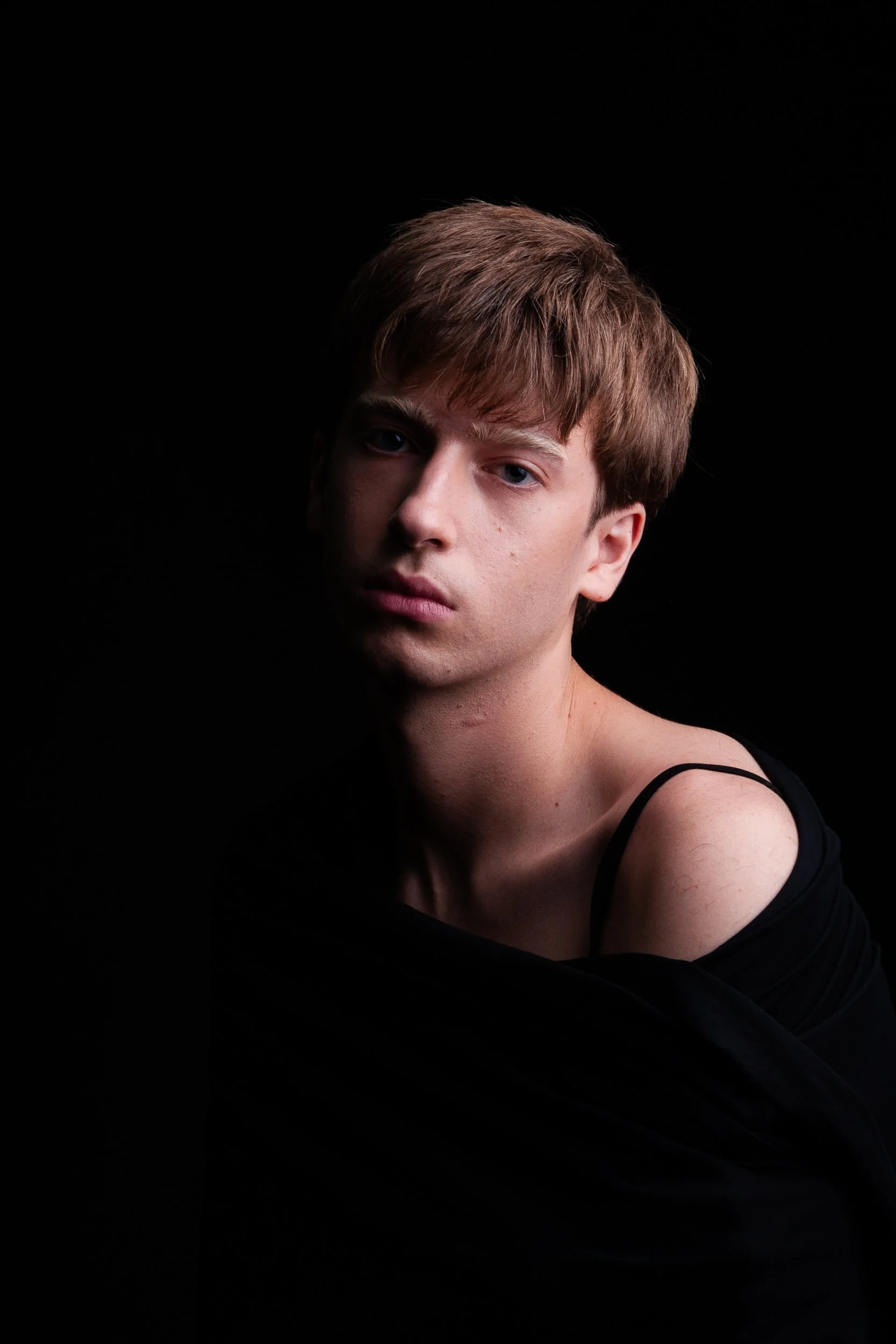 Moody close-up portrait of a young man with brown hair wearing a black shirt against a black background