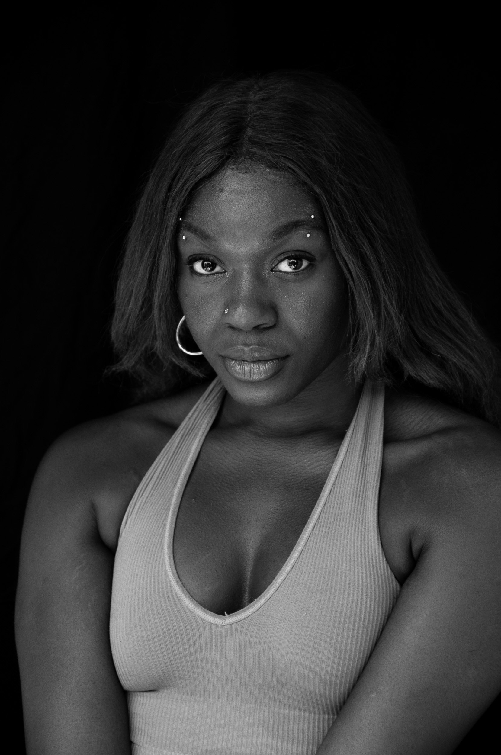 Black-and-white portrait of a woman with long hair, facial piercings, hoop earrings, and a ribbed sleeveless top against a dark background