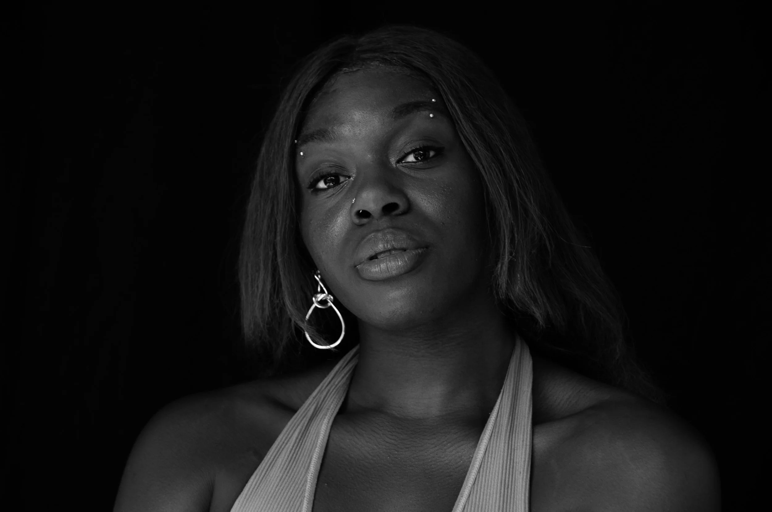 Black-and-white close-up portrait of a woman with long hair, hoop earrings, facial piercings, and dramatic eye makeup against a black background.