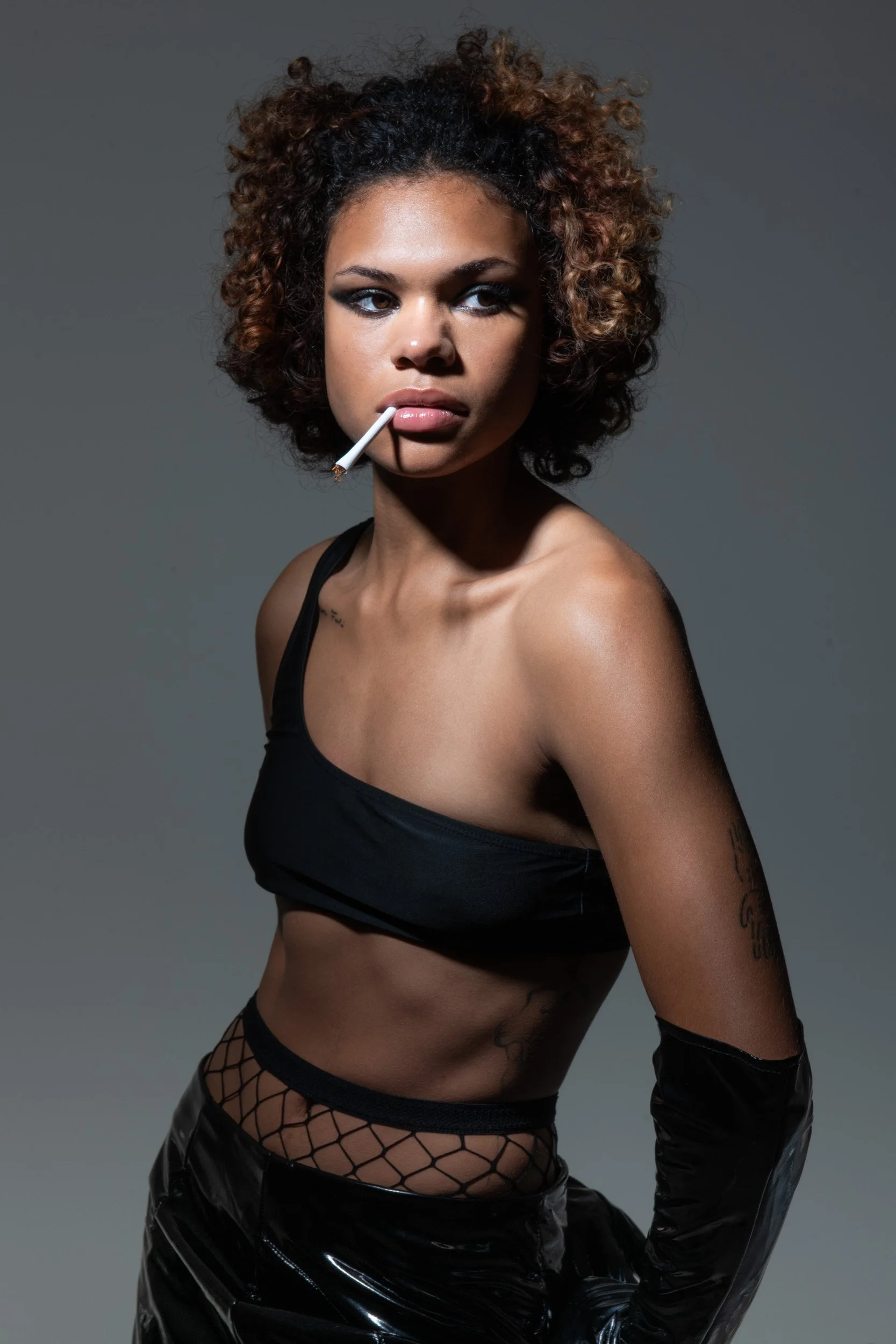 Studio portrait of a curly-haired woman in black clothing holding a lollipop against a gray background