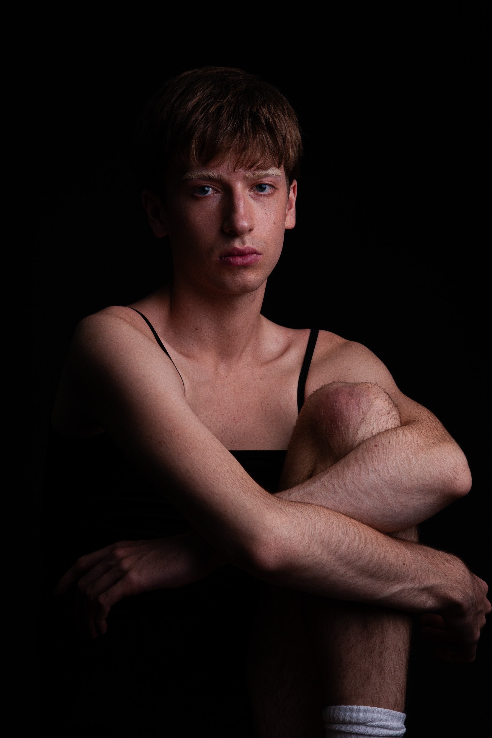 Portrait of a young man with brown hair and blue eyes sitting cross-legged against a black background