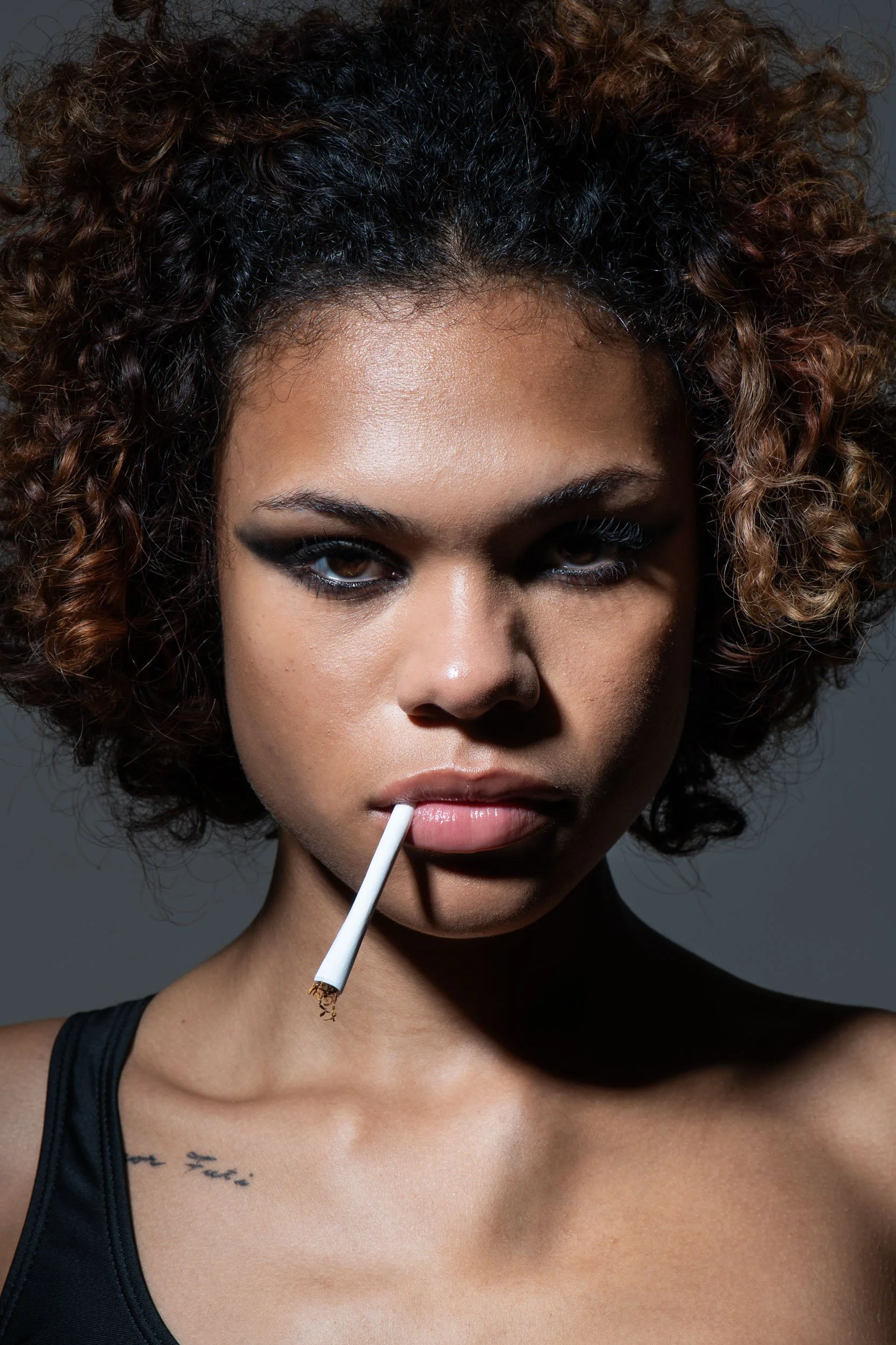 Close-up portrait of a woman with curly hair, dramatic eye makeup, and a cigarette between her lips