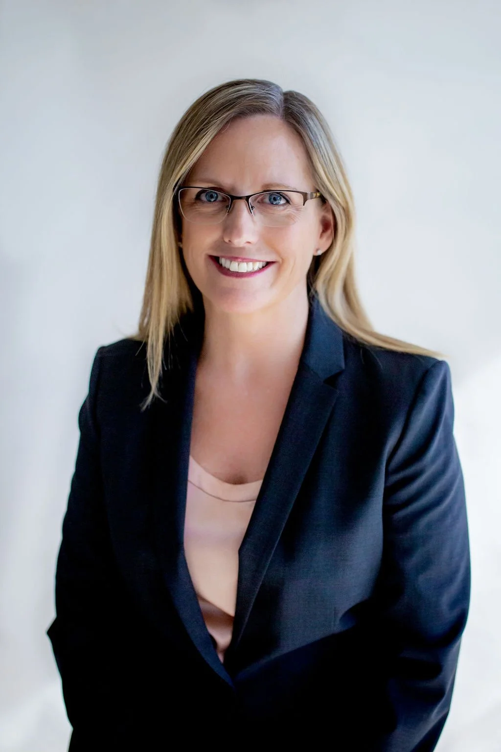 A woman with blonde hair, glasses, and a friendly smile, wearing a dark blazer and light-colored top, standing against a plain wall.