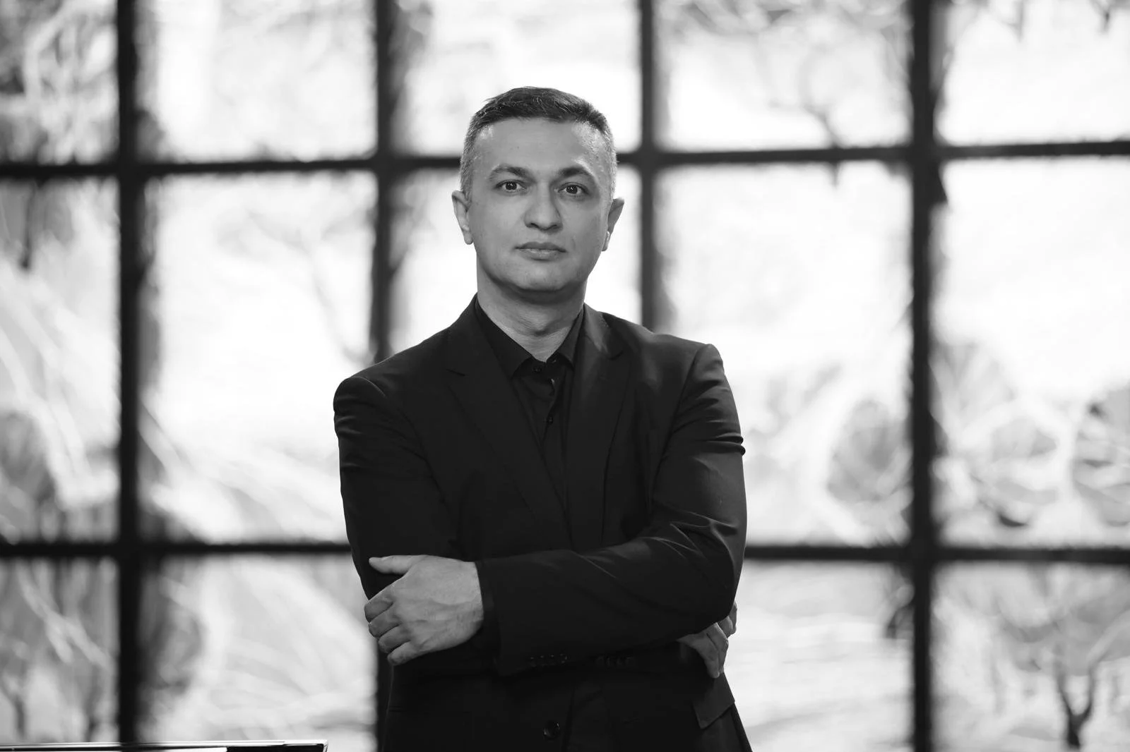 Black-and-white portrait of a man in a dark suit standing with crossed arms in front of large windows.