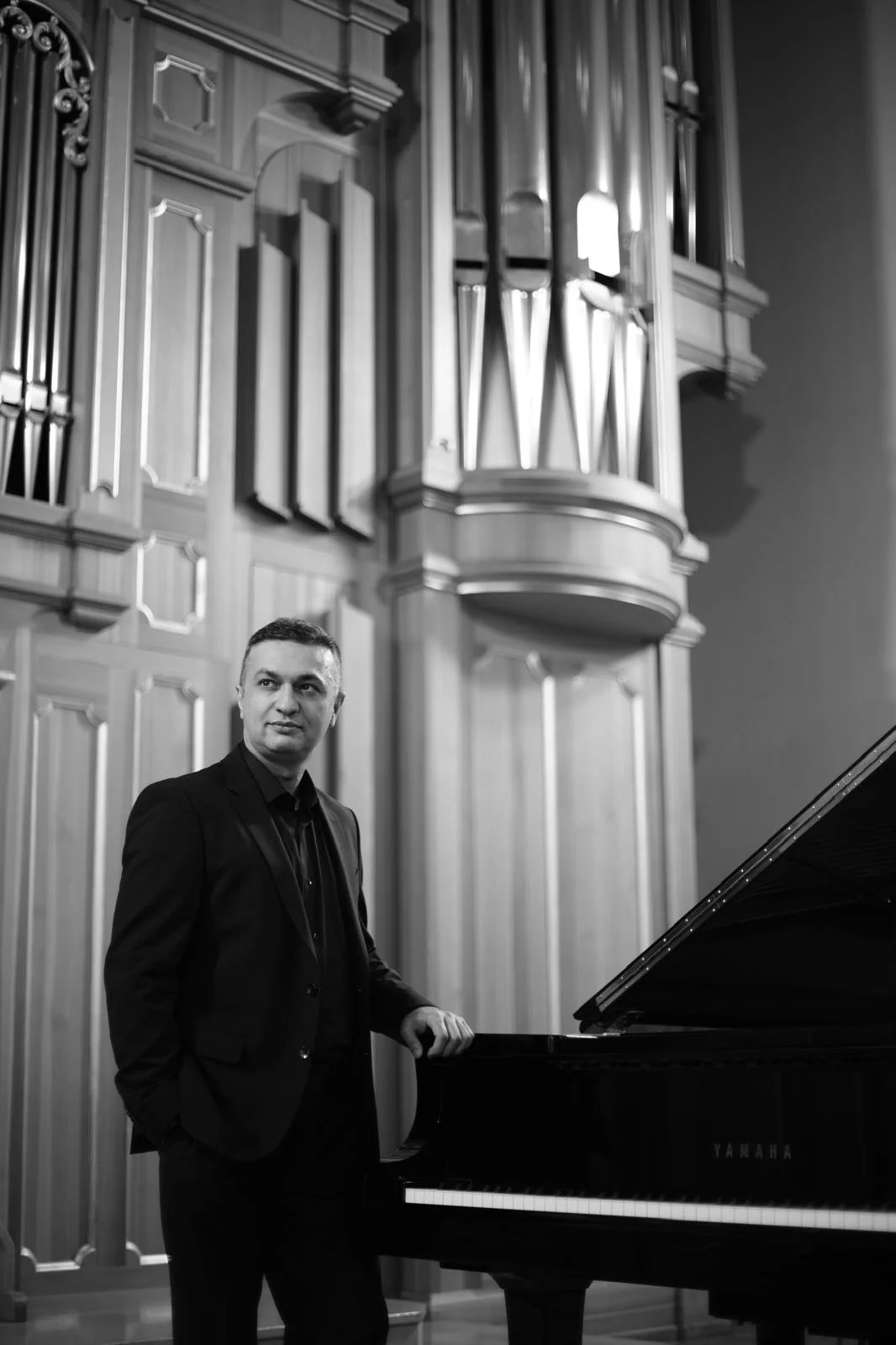 Black-and-white portrait of a man in a suit standing beside a Yamaha grand piano in an ornate concert hall.