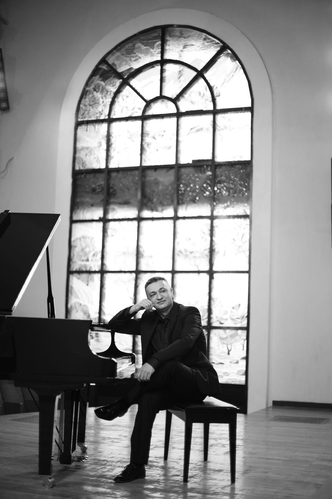 Black-and-white portrait of a man in a suit seated beside a grand piano in front of a tall arched stained-glass window