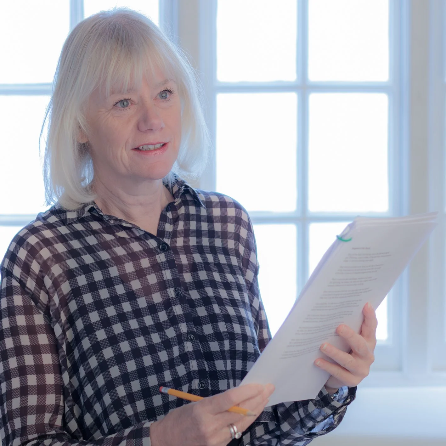 Mature woman in a checkered shirt reviewing documents with a pencil by a bright window