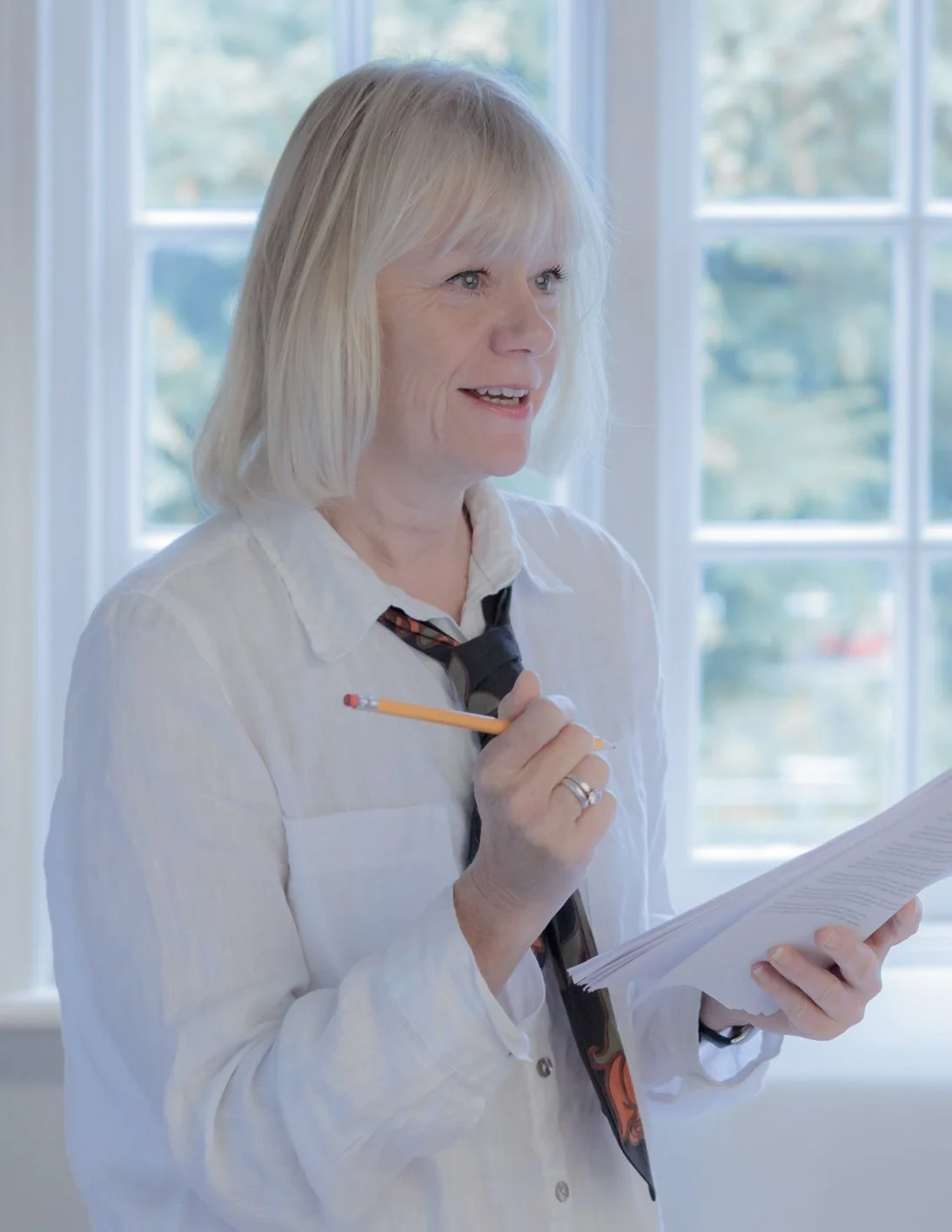 Blonde woman in a white shirt holding a pencil and documents near a window