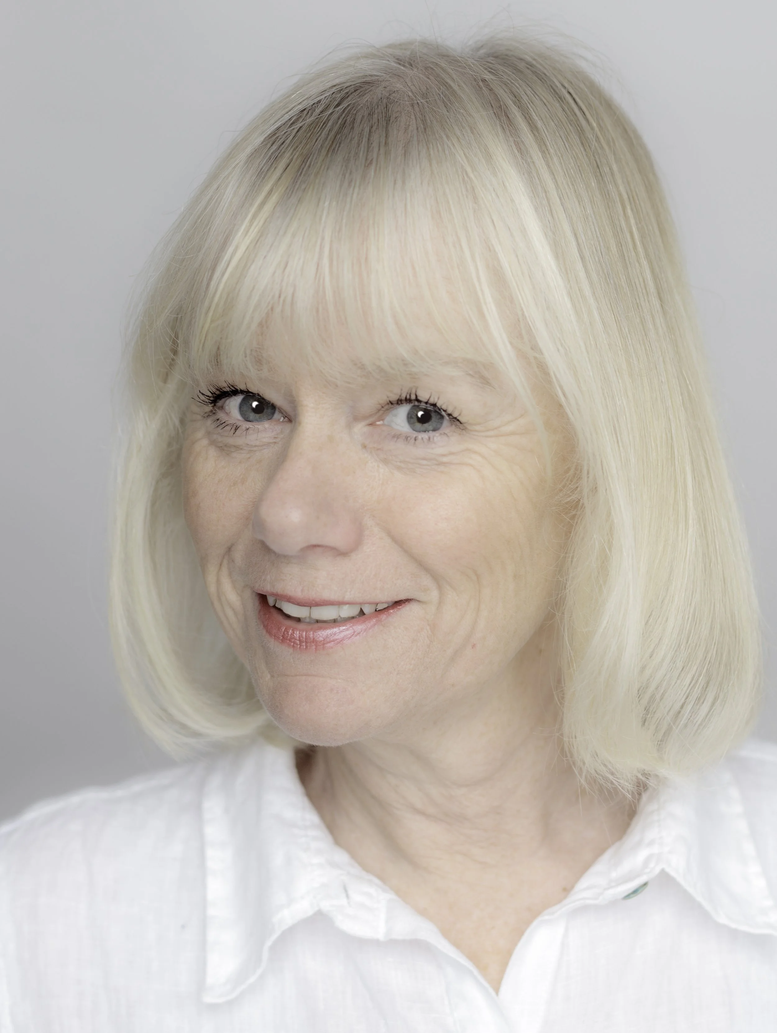 Close-up portrait of a smiling older woman with blond hair and blue eyes wearing a white shirt