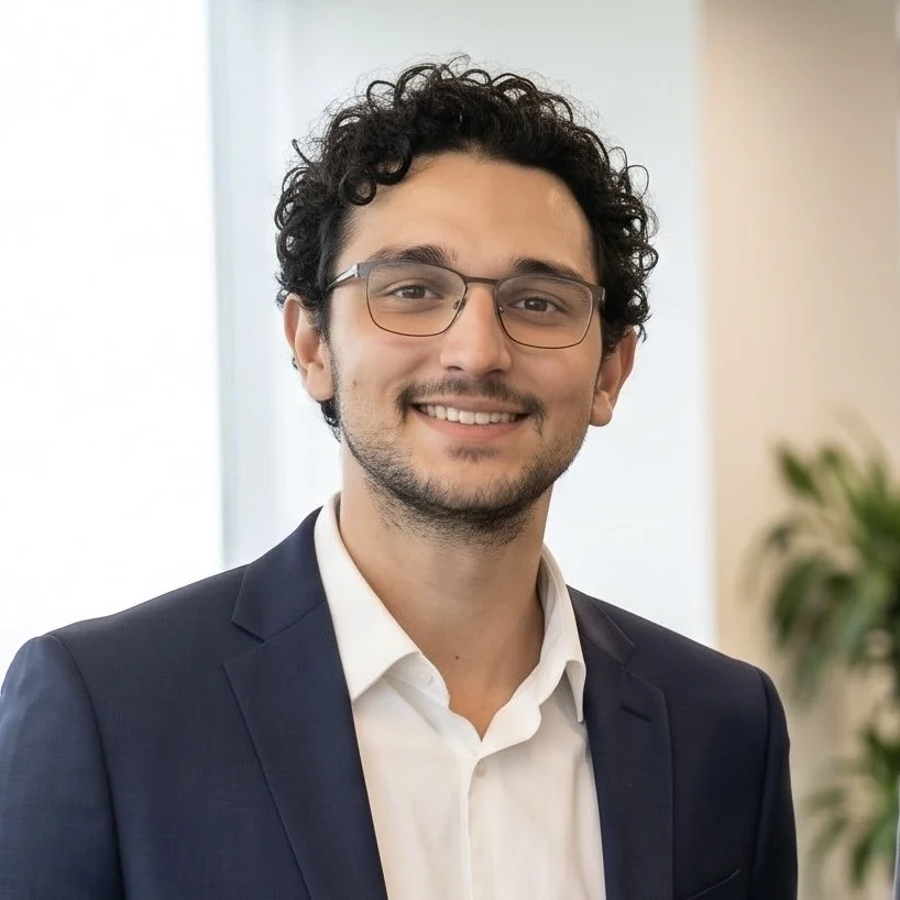Smiling man with curly hair and glasses wearing a navy suit and white shirt