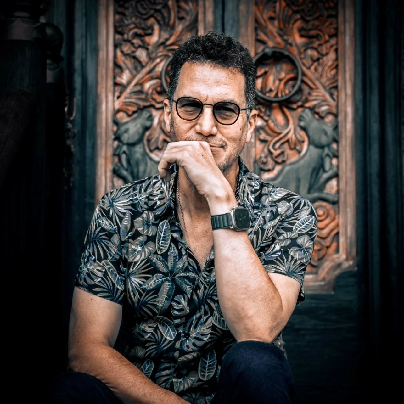 Steven Shulem in sunglasses and patterned shirt sitting in front of ornate wooden door, wearing a smartwatch.