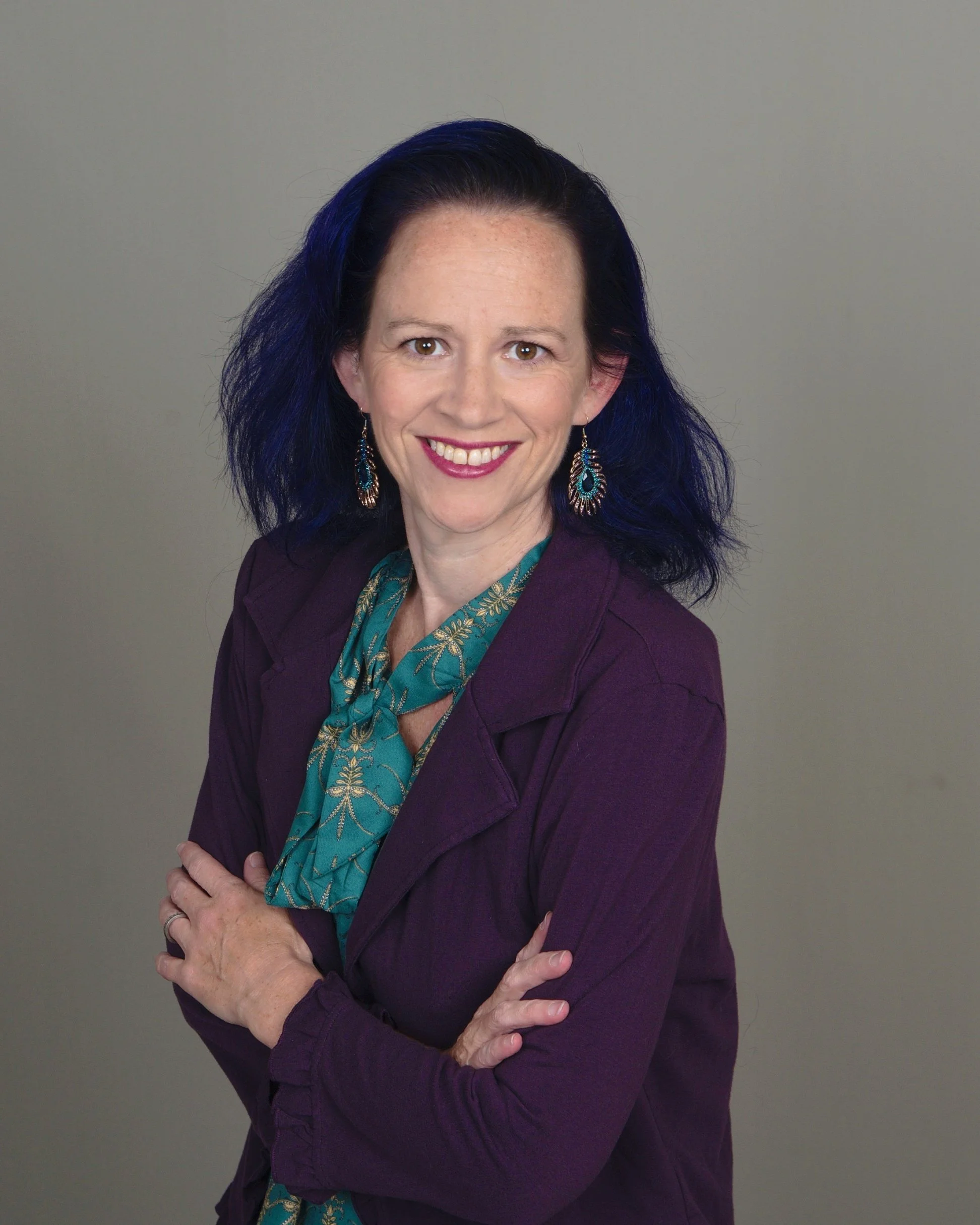 Smiling woman with dark blue hair wearing a teal blouse with gold pattern, purple blazer, and large turquoise earrings, standing against plain gray background.