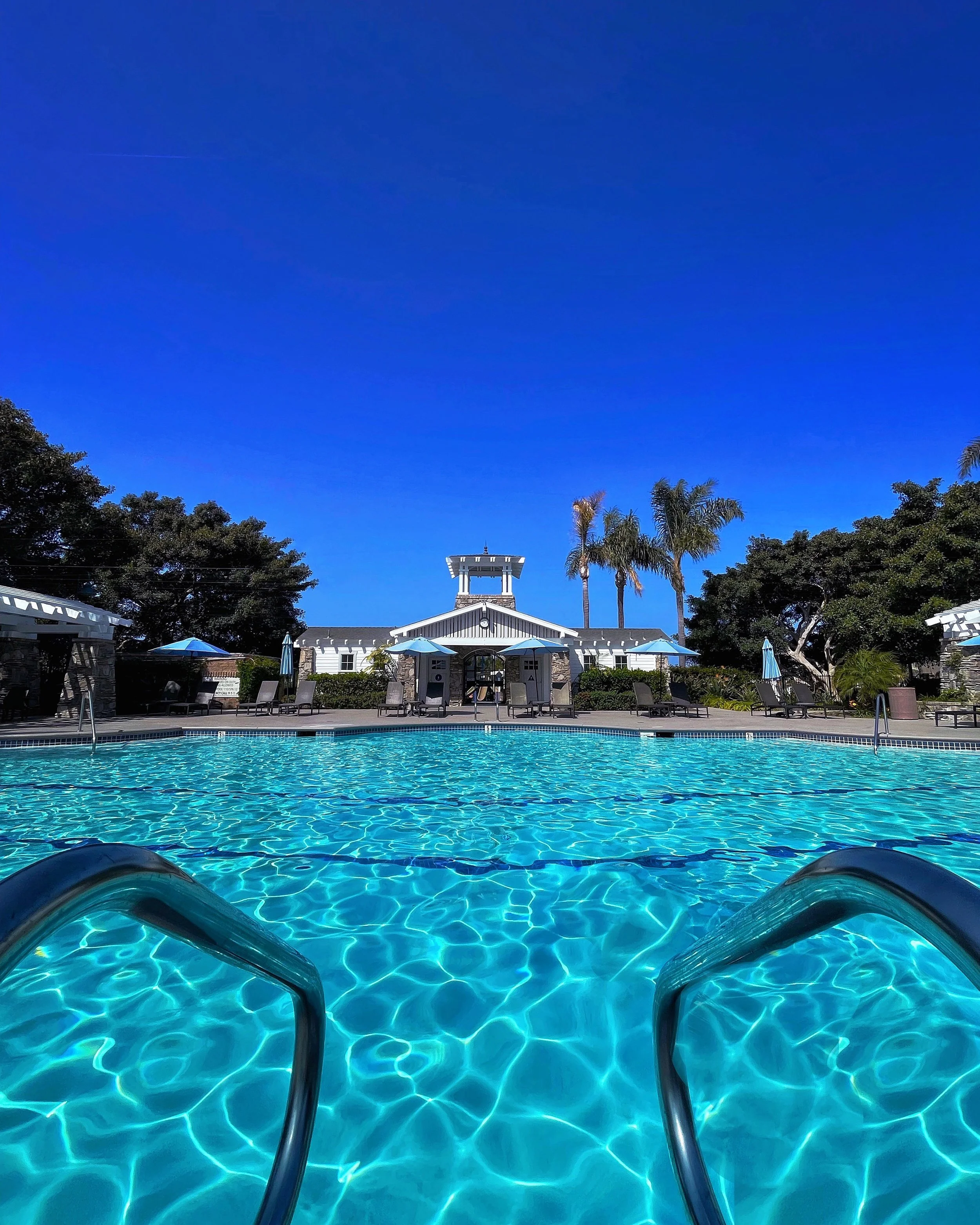 View of a Commercial Aquatics swimming pool San Diego account of ours with clear blue water, pool ladder in the foreground, poolside lounge chairs with blue umbrellas, a white community building with a clock and bell tower, surrounded by trees.