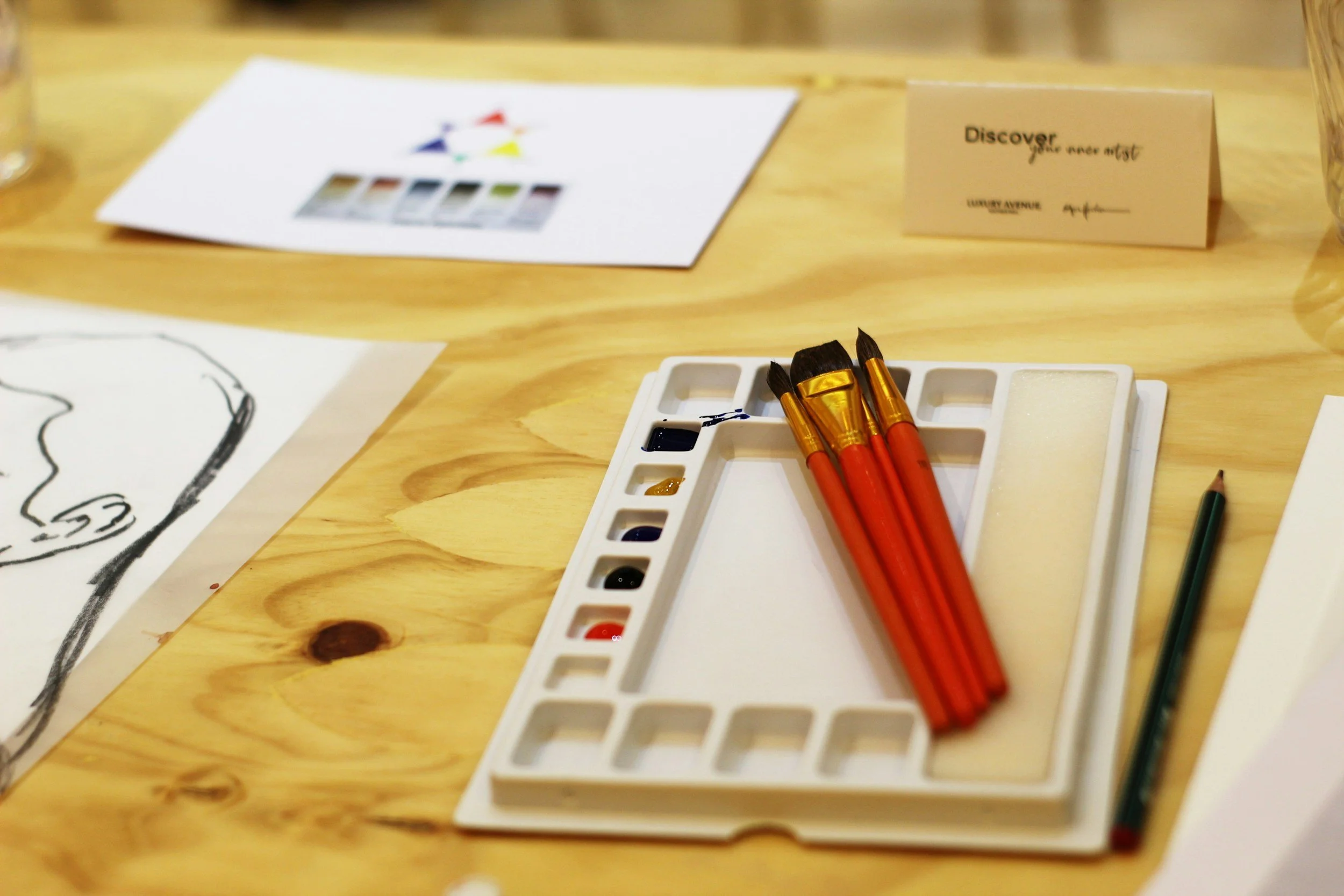 Paleta de acuarelas con pinceles rojos y lápiz sobre una mesa de madera, junto a dibujos y una tarjeta.