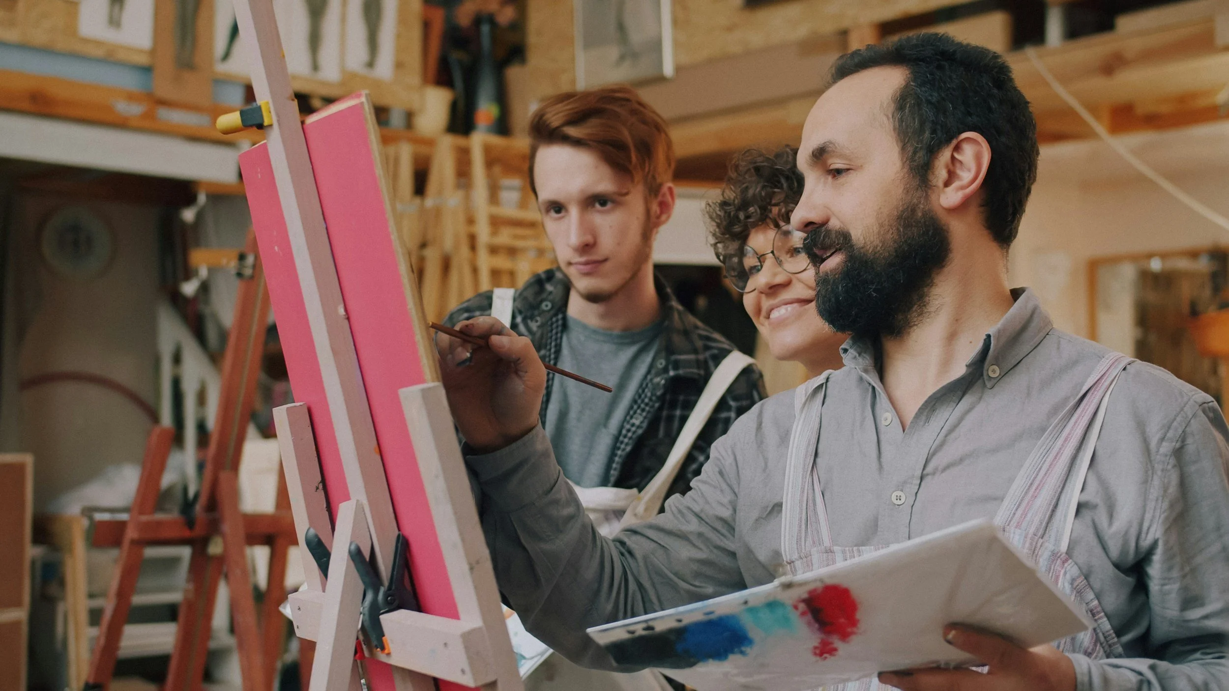 Tres artistas pintan juntos en un estudio de arte frente a un caballete con un lienzo rosa.