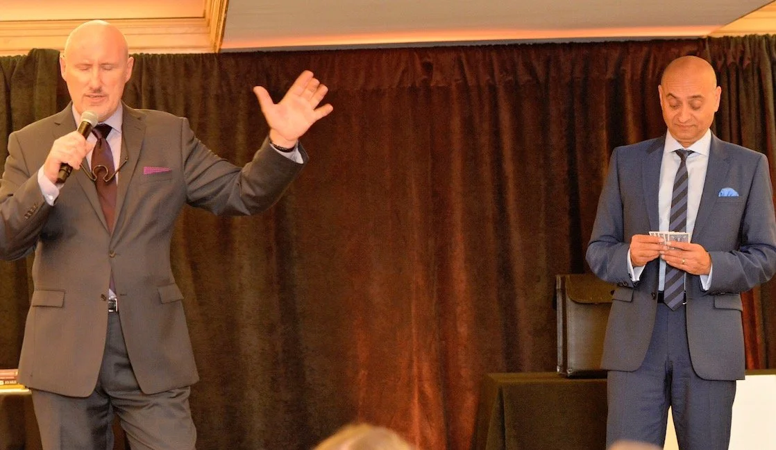 Two men in suits on a stage, one speaking into a microphone while the other holds a card.