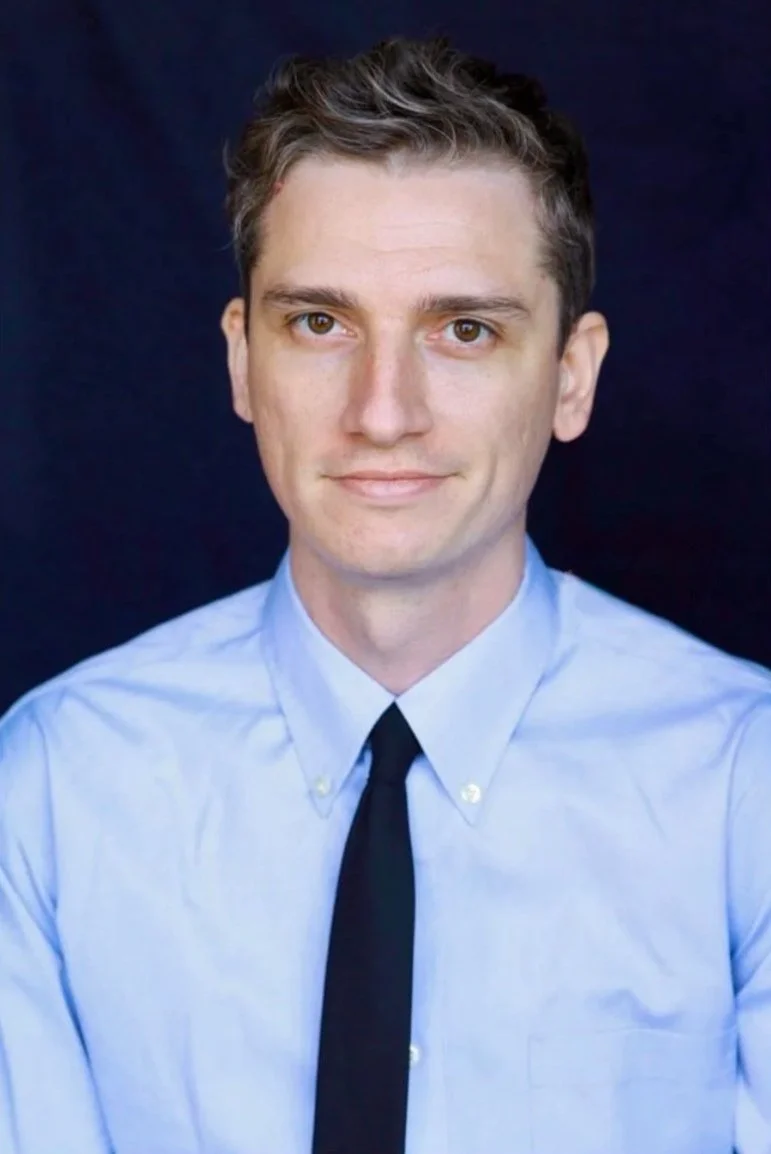 A young man with brown hair wearing a light blue dress shirt and black tie against a dark background.