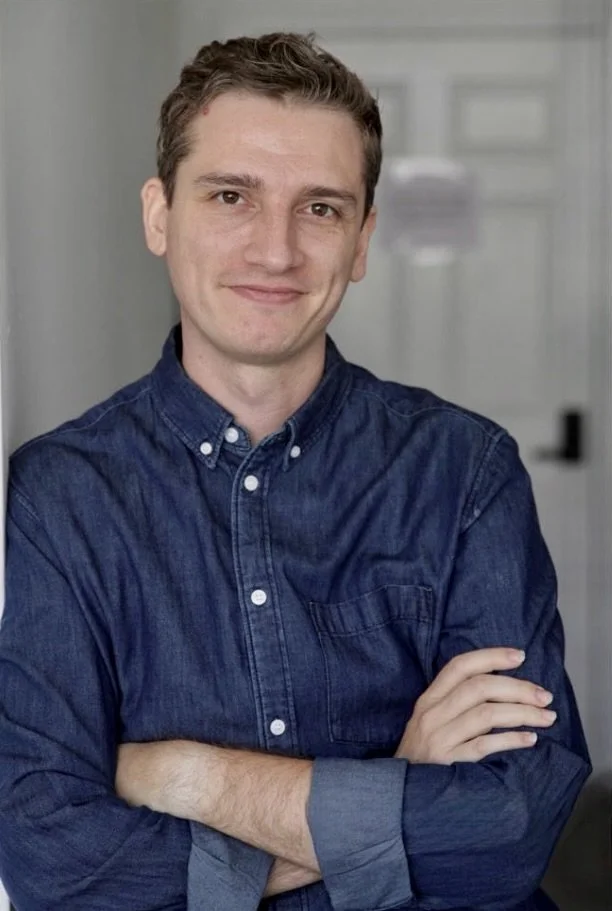 A young man with short brown hair and a slight smile, wearing a dark blue button-up shirt with rolled-up sleeves, standing with his arms crossed in an indoor setting.