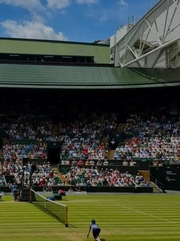 A tennis court with a player preparing to serve, surrounded by a large crowd watching from grandstands under a blue sky with some clouds.