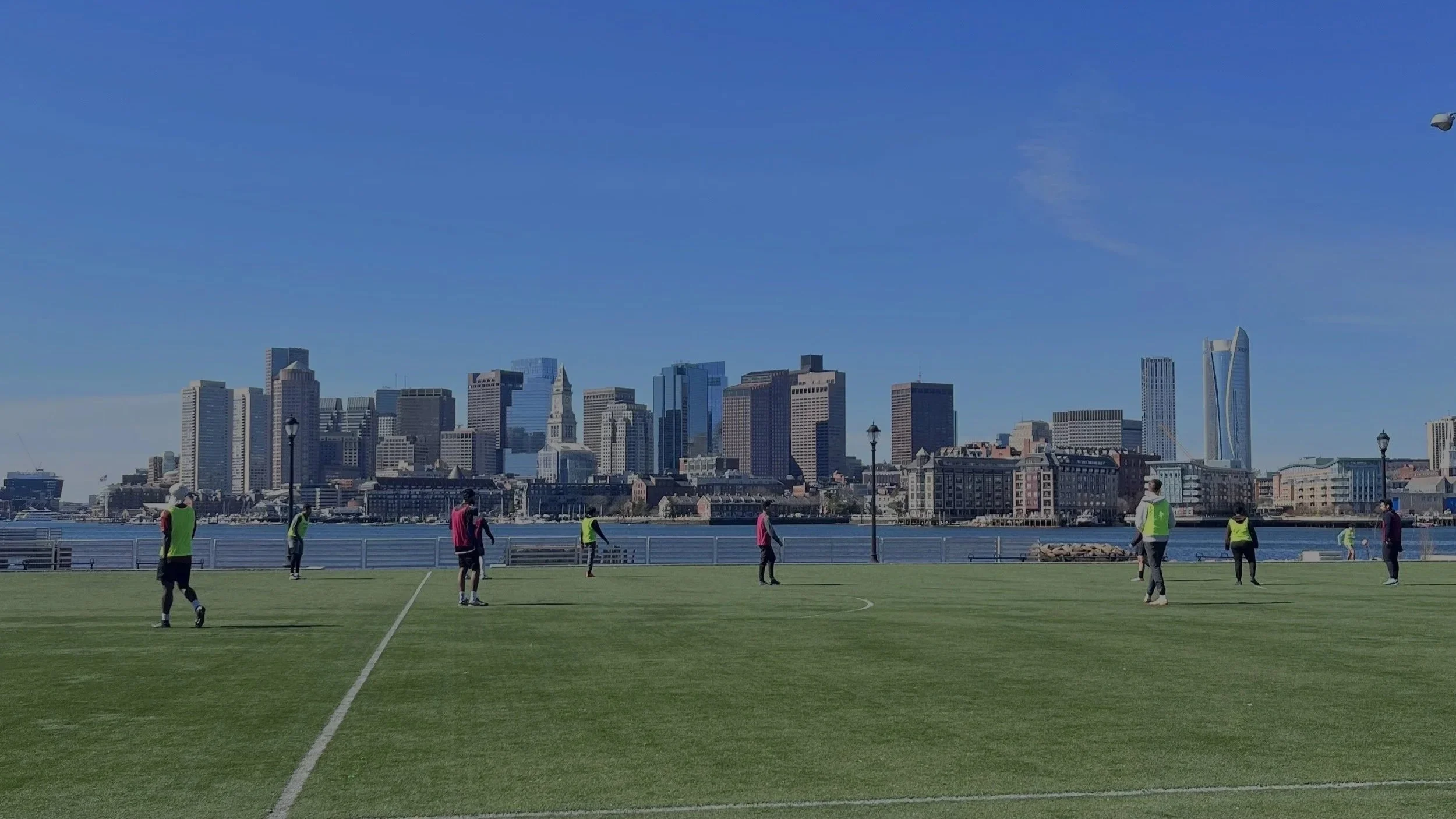 People playing soccer on a green field with a city skyline of tall buildings and skyscrapers in the background under a blue sky.