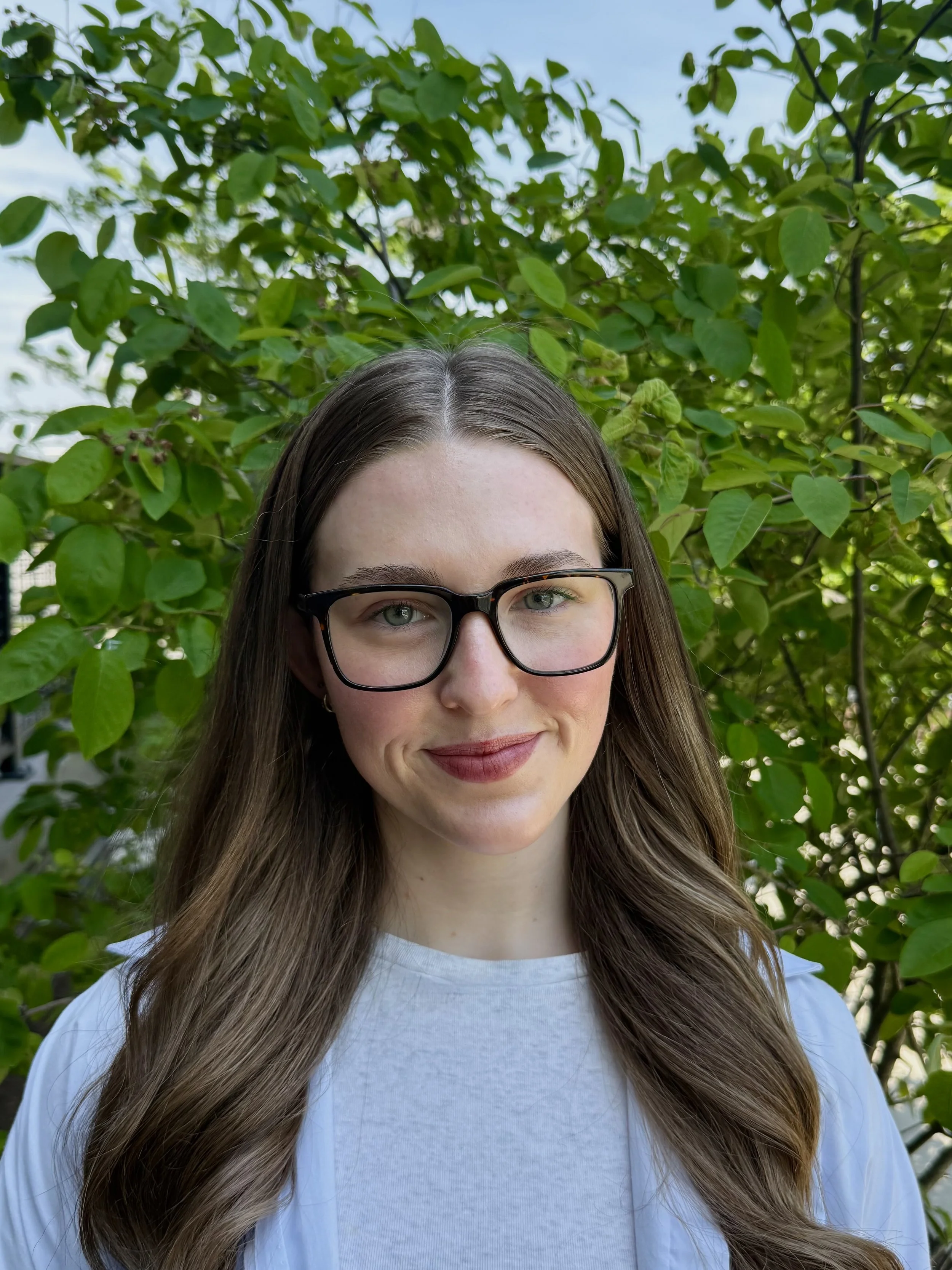 Close-up of a woman with long brown hair, glasses, and a light-colored shirt, standing outdoors in front of green leafy plants and a partly cloudy sky.