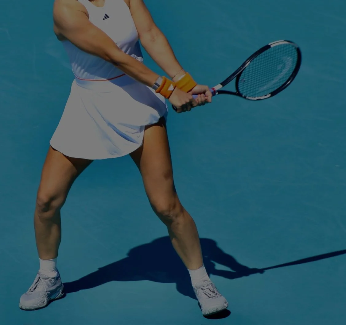 A female tennis player is preparing to hit a tennis ball on a blue court, holding a tennis racket with both hands and wearing a white athletic outfit.