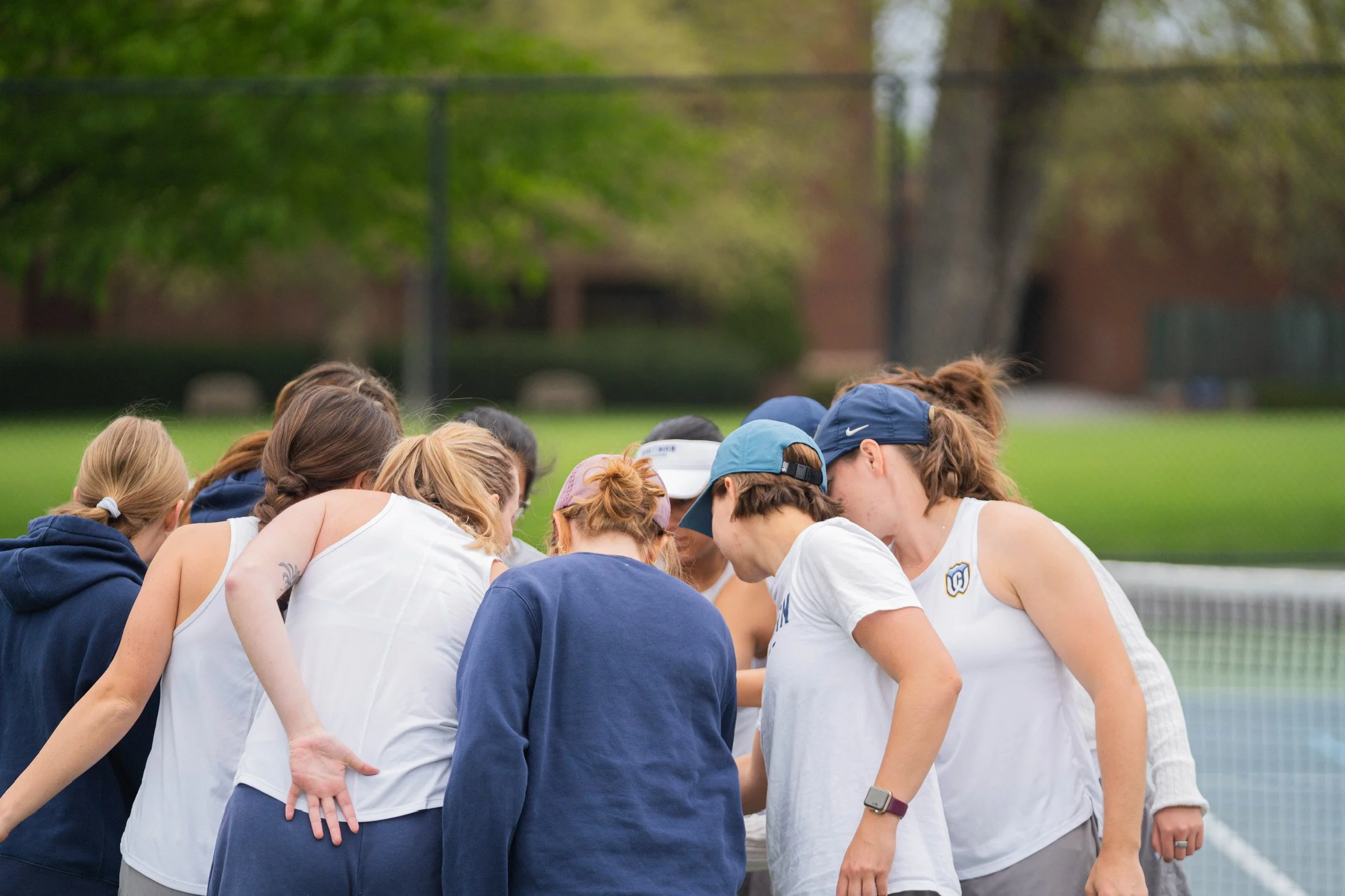 A group of individuals huddled together on an outdoor tennis court, possibly in a team huddle or discussion. They are wearing sports clothing, including white shirts and various hats. Green trees and a chain-link fence are visible in the background.