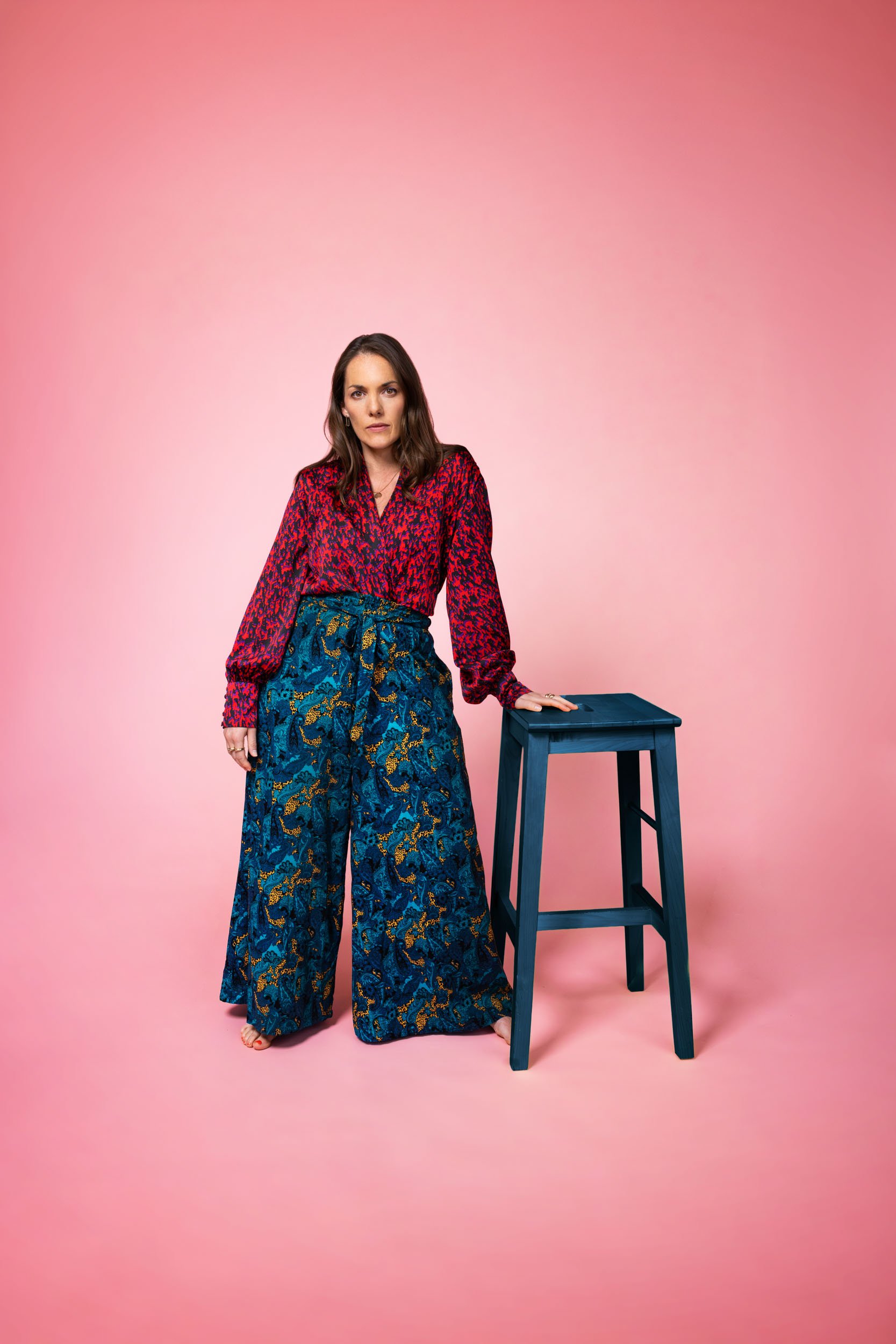 Maria Finkelmeier standing up, leaning on a stool, on a pink background