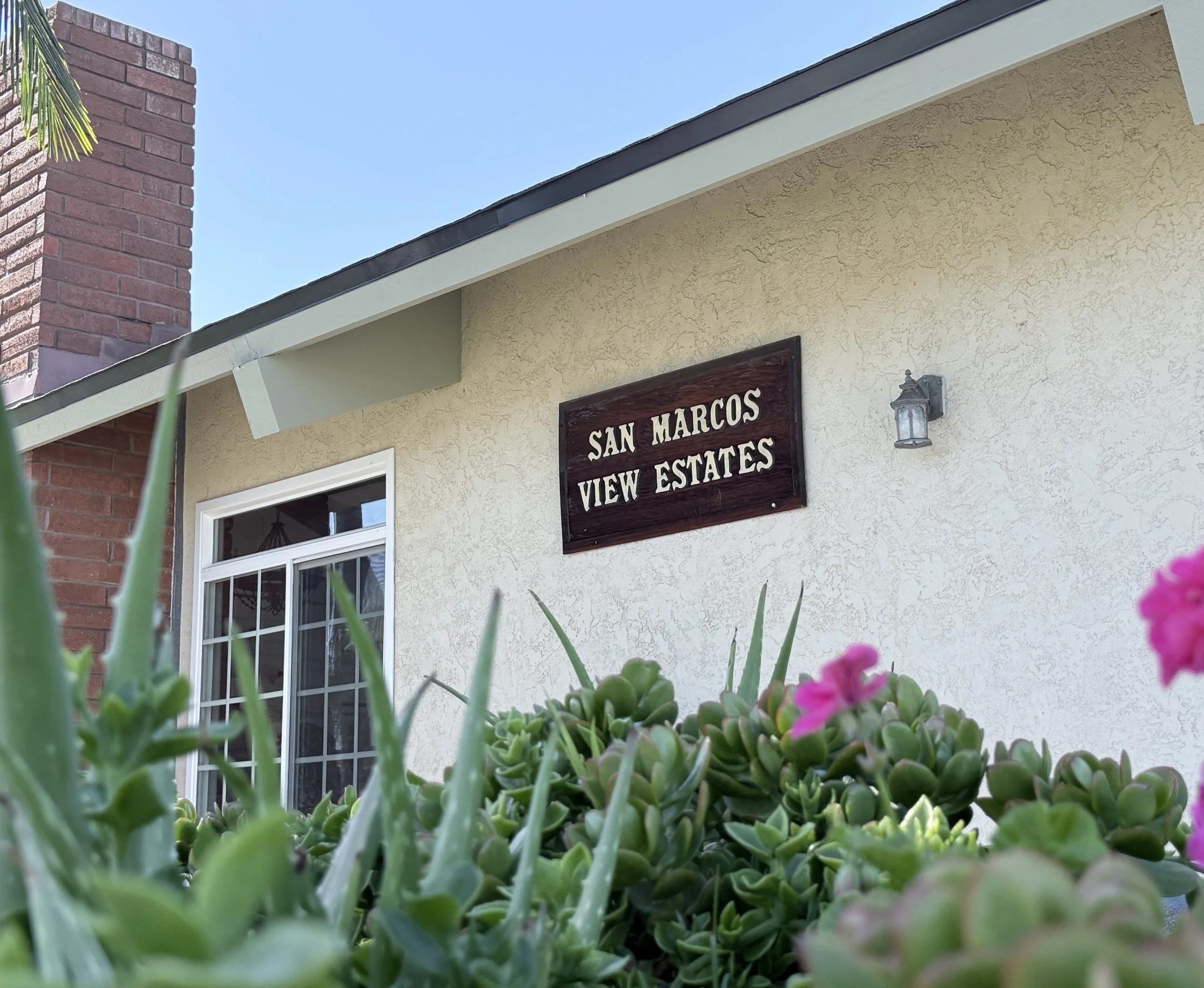 San Marcos View Estates sign on the clubhouse wall, behind succulents and pink geraniums.