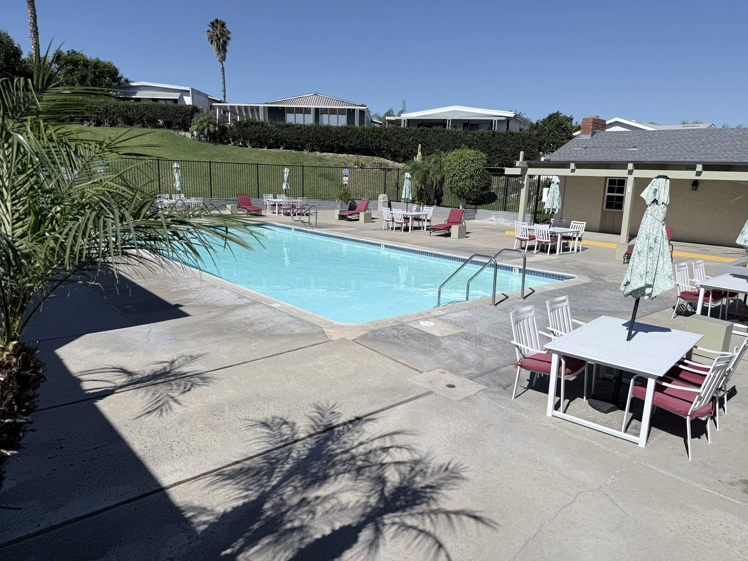 Community pool with patio tables, lounge chairs and palm trees, with hillside homes in the background.