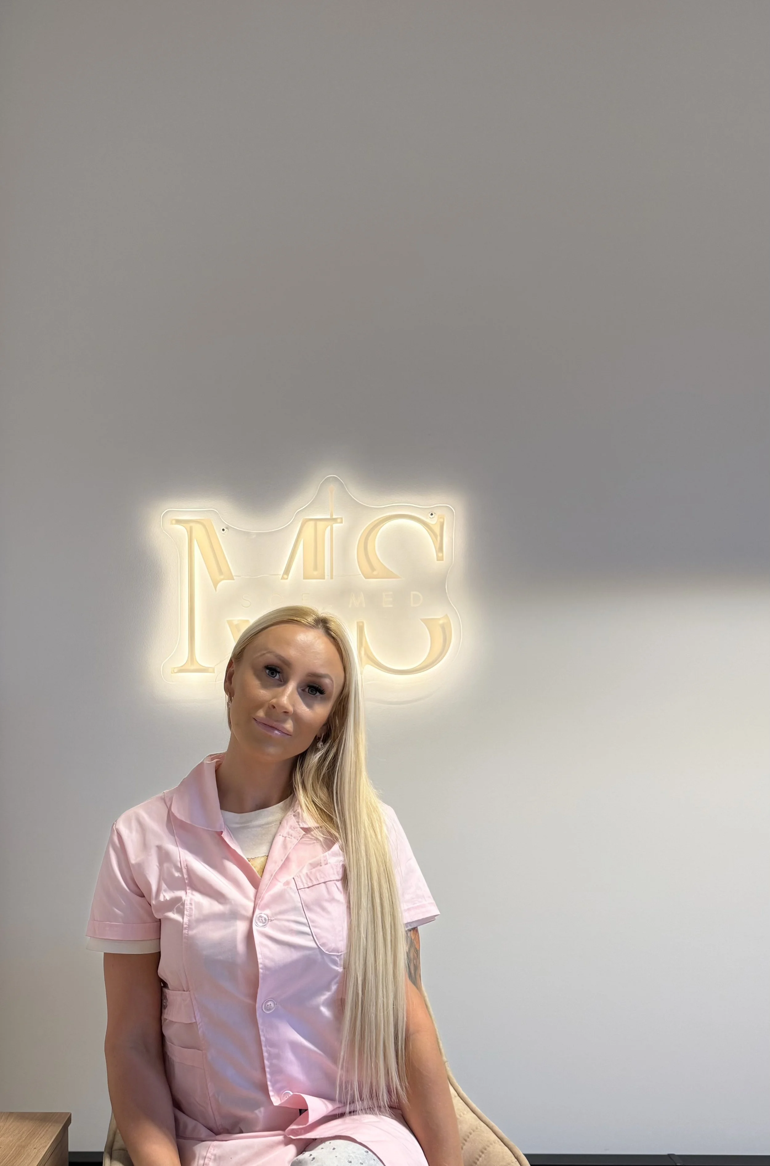 Blonde woman in pink medical scrubs seated beneath an illuminated aesthetic medicine clinic sign