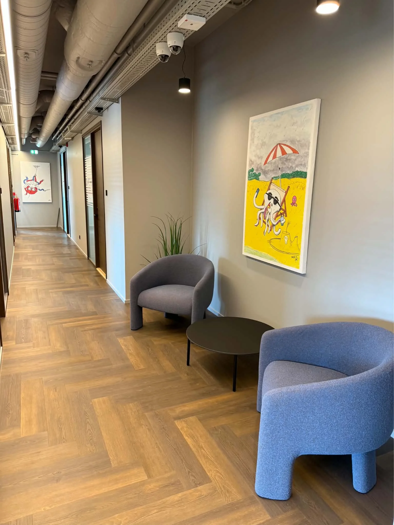 Modern office hallway lounge with blue and gray chairs, round black table, colorful artwork, exposed ceiling ducts, and security cameras