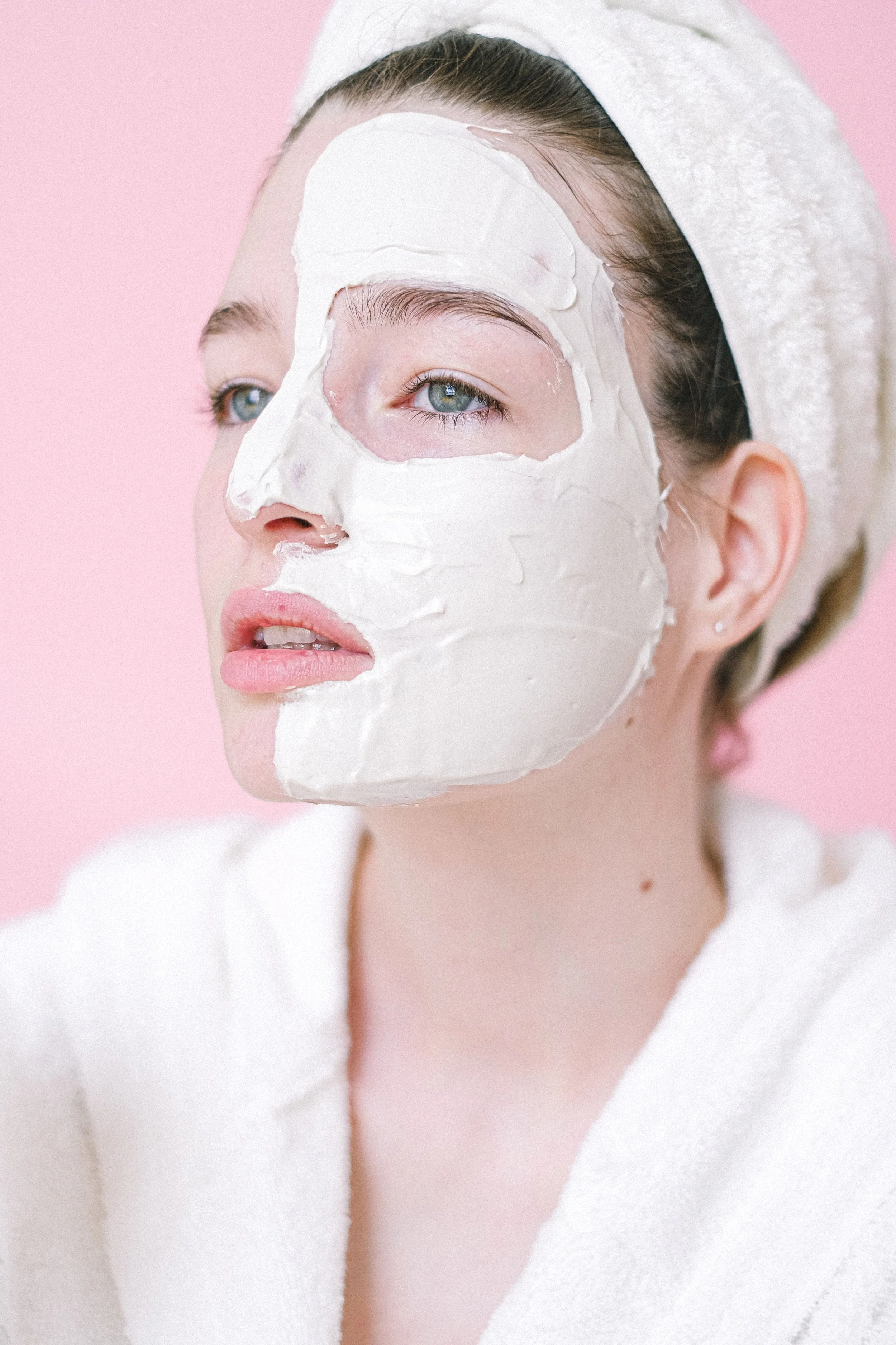 Close-up of a woman wearing a white facial sheet mask against a pink background