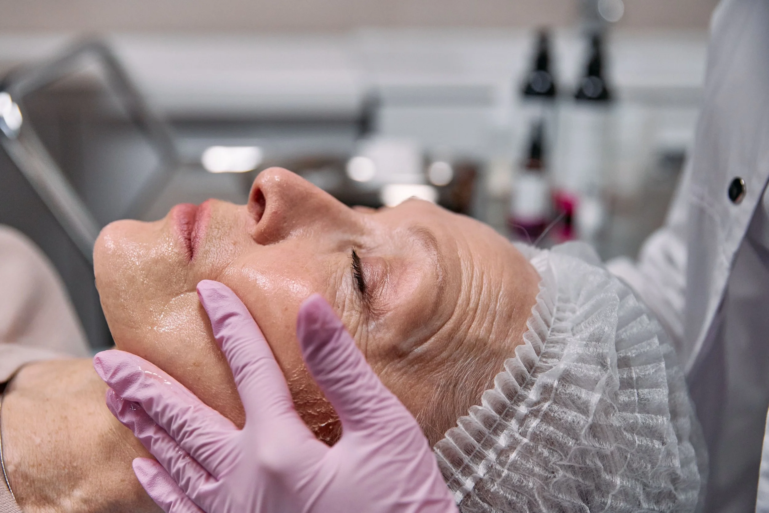 Client receiving a facial skincare treatment from a practitioner wearing pink gloves and a disposable hairnet
