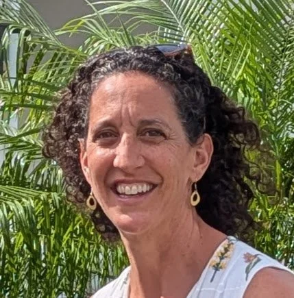 Woman with curly hair smiles outdoors with green tropical plants in the background.