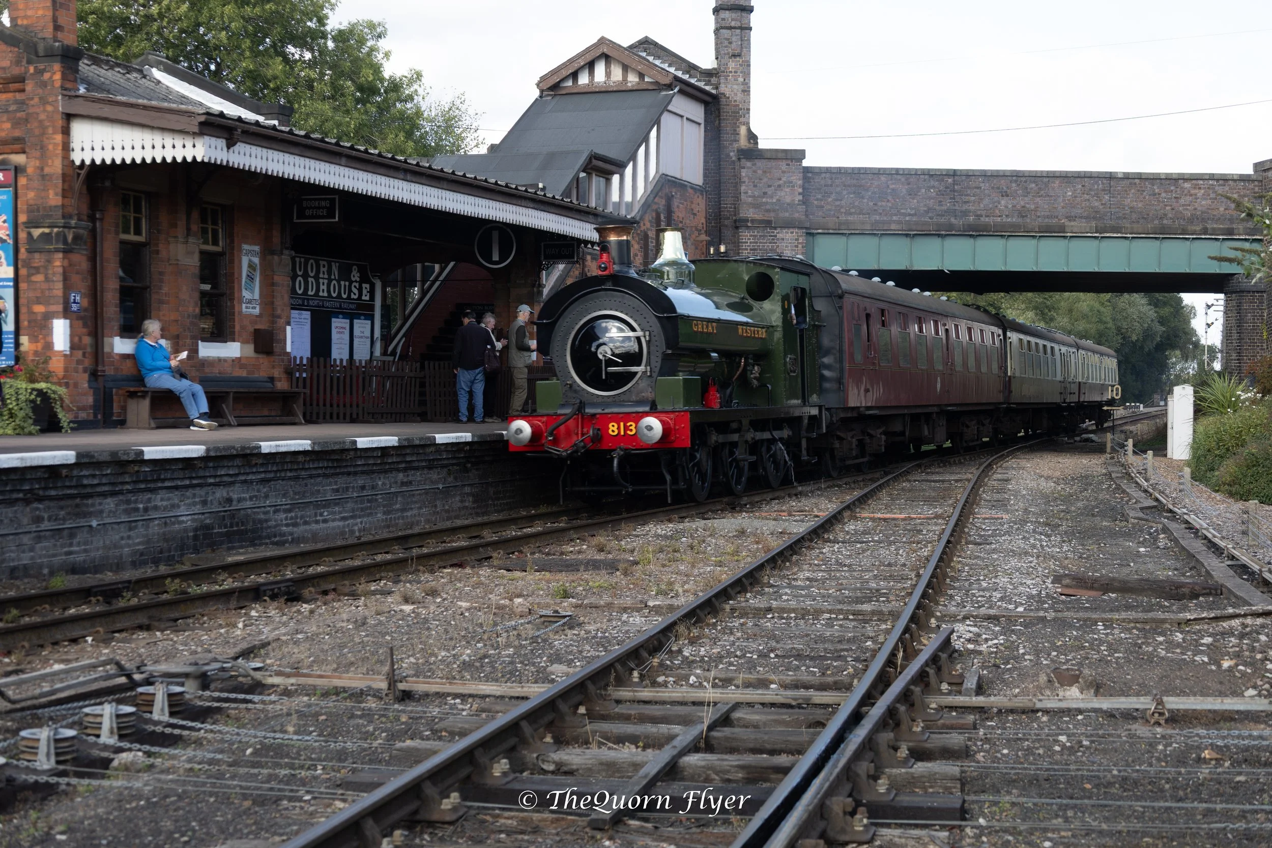 A vintage steam train arriving at a historic station with a brick building, people waiting, and tracks in the foreground.