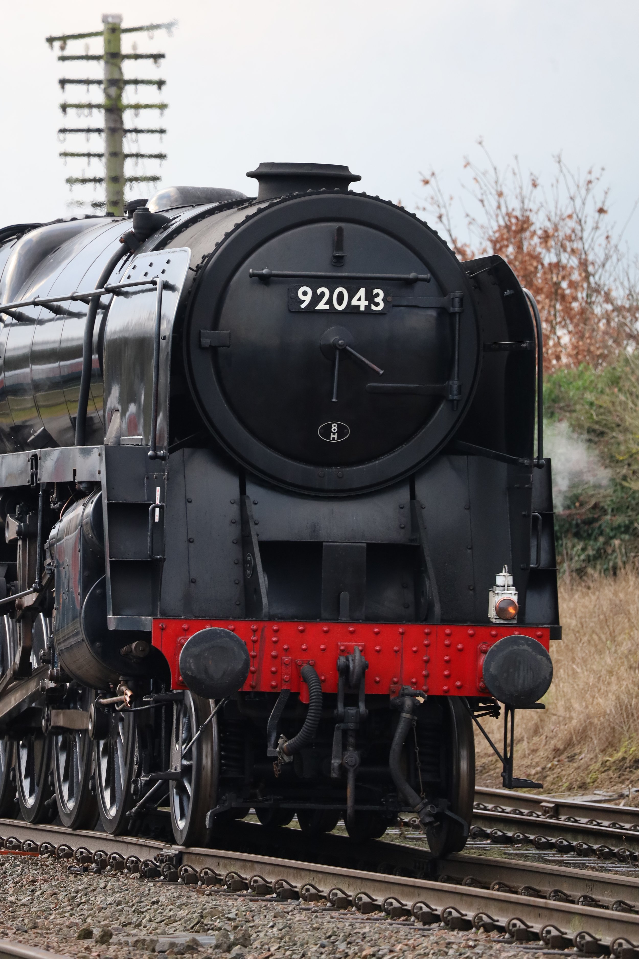 A black steam locomotive with the number 92043 on the front, on train tracks outdoors, with a background of trees and an electrical pole.