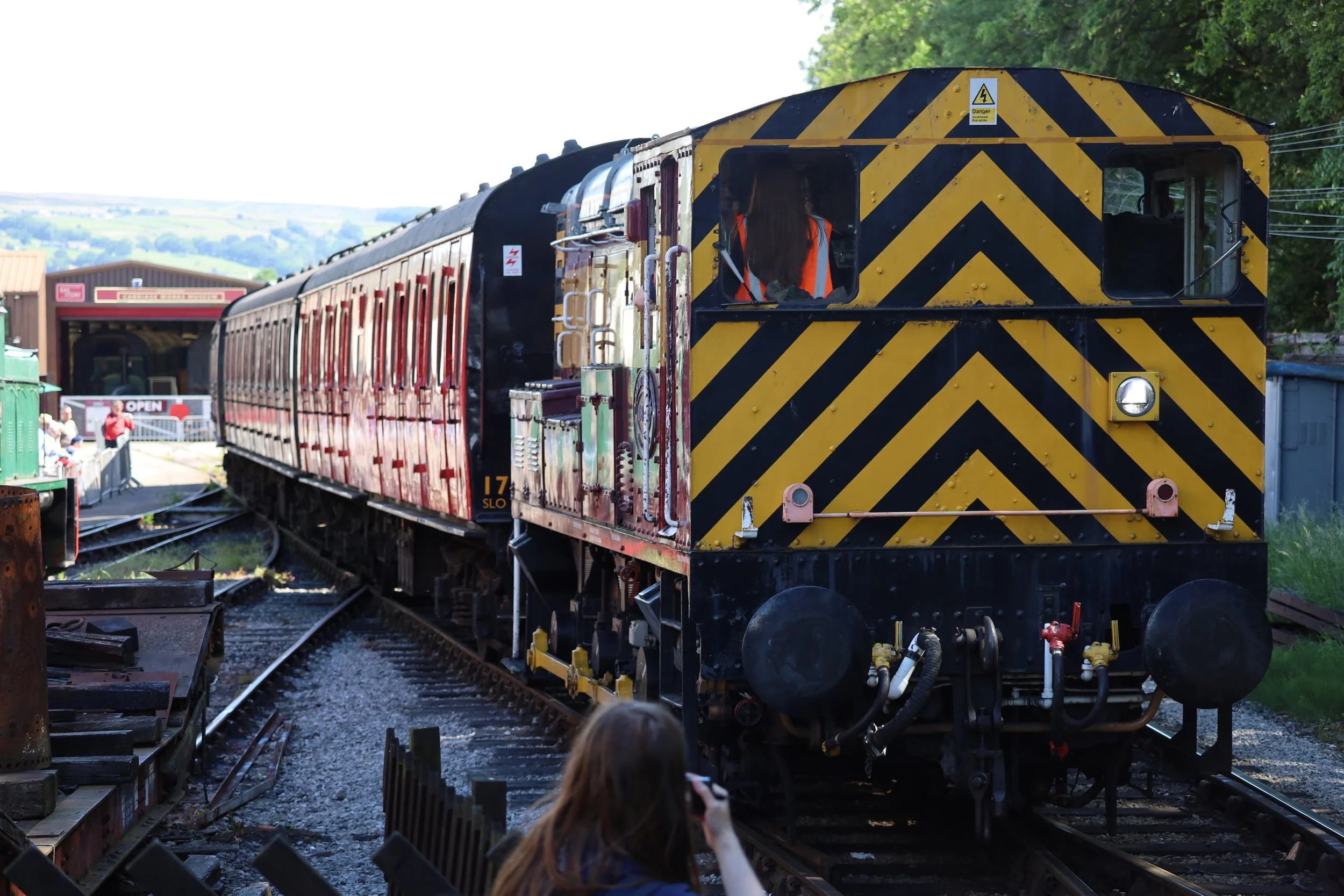 A train engine with black and yellow diagonal stripes at the front, pulling red passenger cars on railway tracks. A person with long hair and an orange vest is inside the train, and a person with a camera is sitting outside near the tracks. There are trees in the background and a building with a sign that says 'Open'.