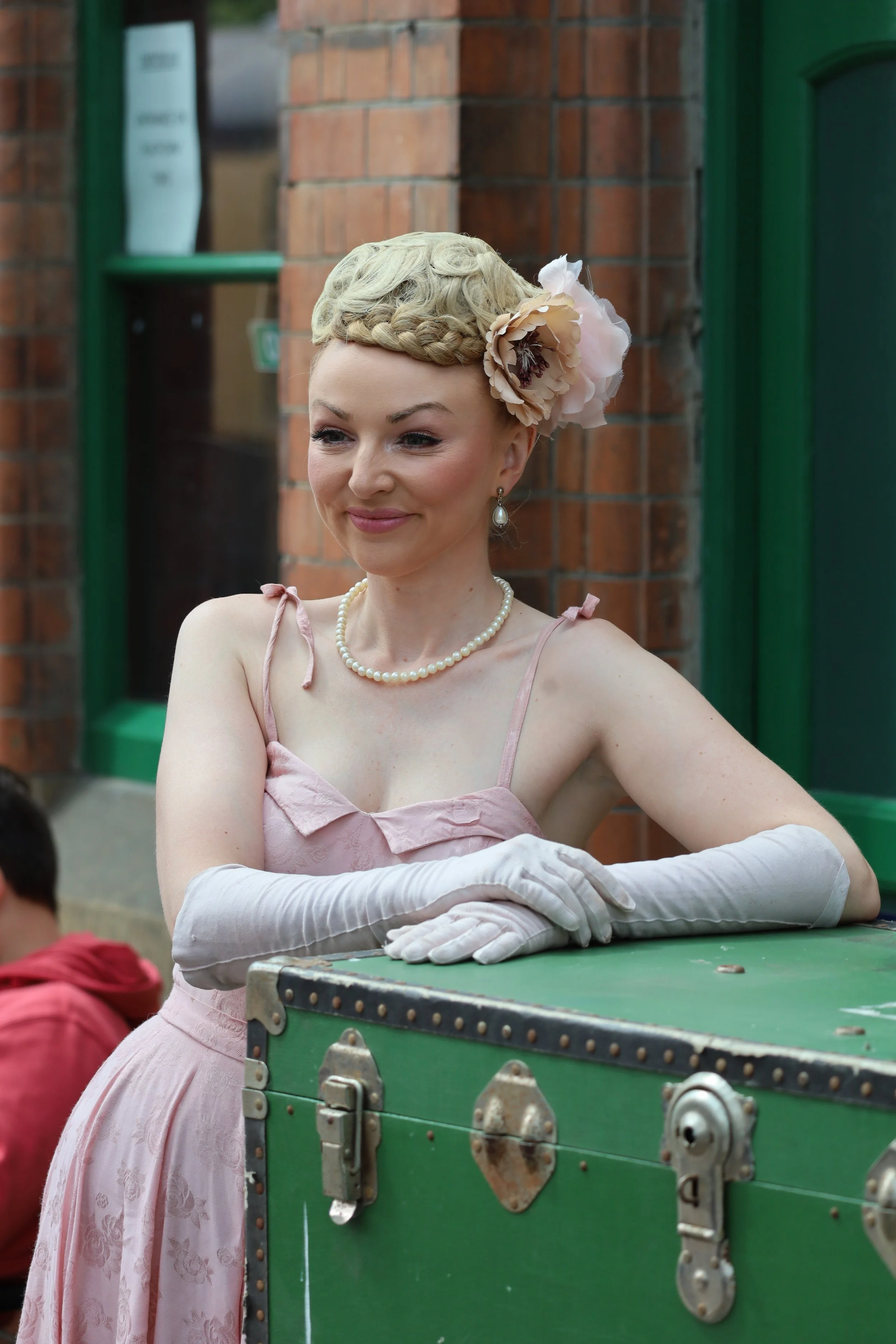 A woman dressed in a vintage pink dress with lace details, wearing white gloves and a pearl necklace, standing next to a green vintage trunk. Her hair is styled in an elaborate updo with a large flower accessory. The background features brick walls and green window frames, indicating an outdoor or street scene.