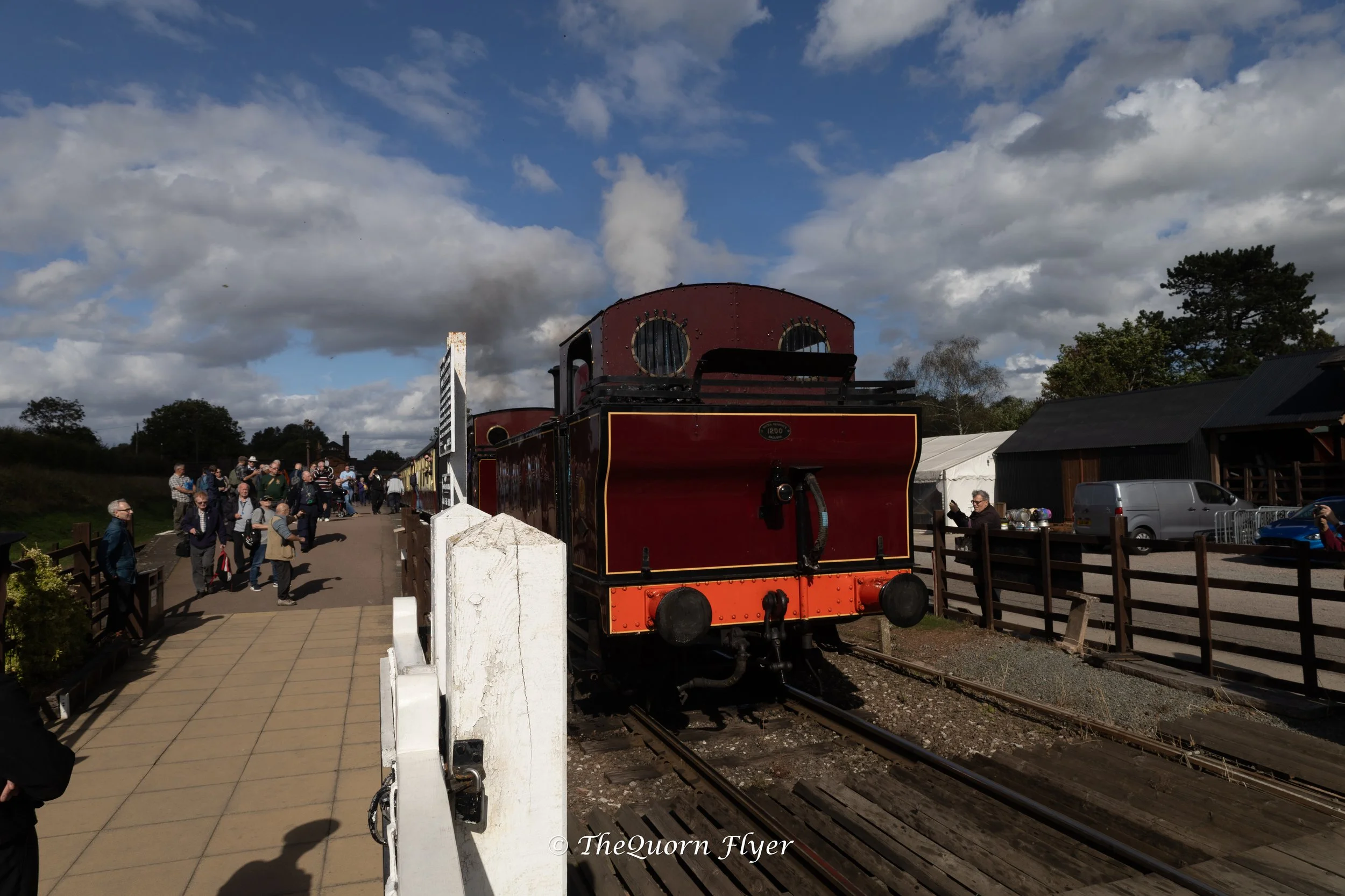 A vintage red steam train with black accents on train tracks, surrounded by a crowd of people at a railway station on a partly cloudy day.
