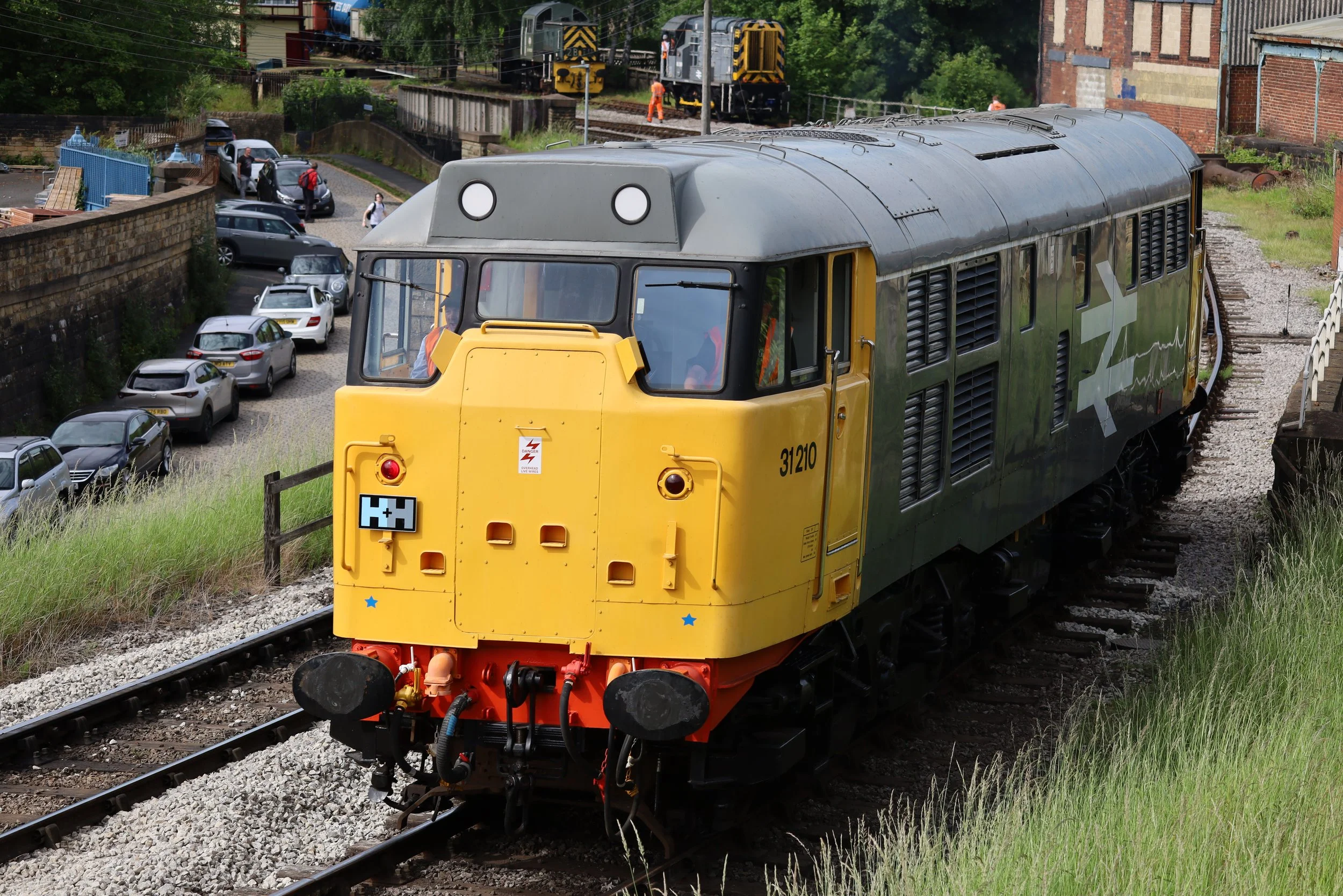 A yellow train locomotive on railway tracks with a gray top and black vents, heading through a semi-urban area with parked cars on one side and a brick building on the other. In the background, there are two dark-colored train cars on an elevated train track, and a train worker in orange safety clothing is near the tracks.