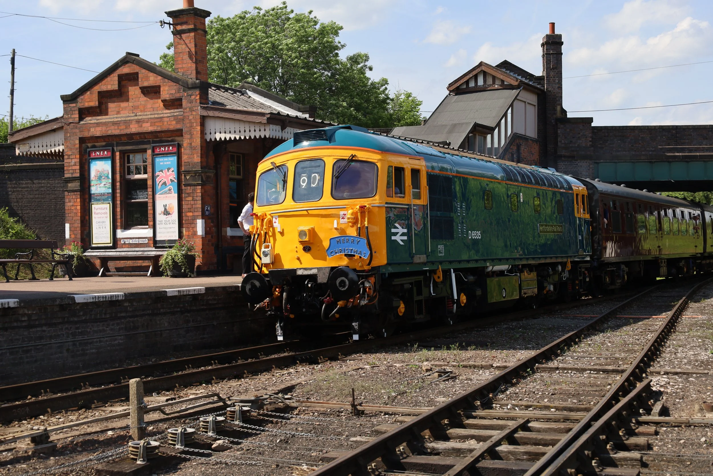 A vintage train with a Christmas decoration on the front is stopped at a small brick train station. The station has a sign that says 'LNER' and a mural with a Union Jack flag. The scene is outdoors with trees and a blue sky.