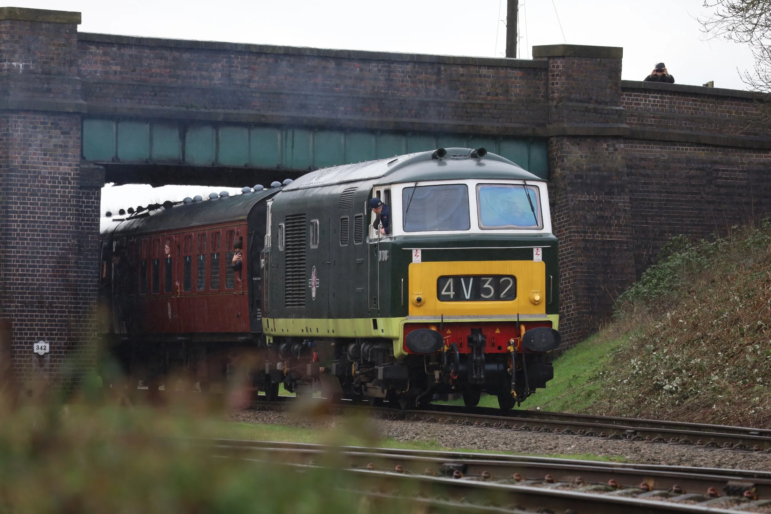 A vintage train with a green and yellow front is passing under a brick bridge. A person is leaning out of the window, and another person is taking a photo from the bridge side.