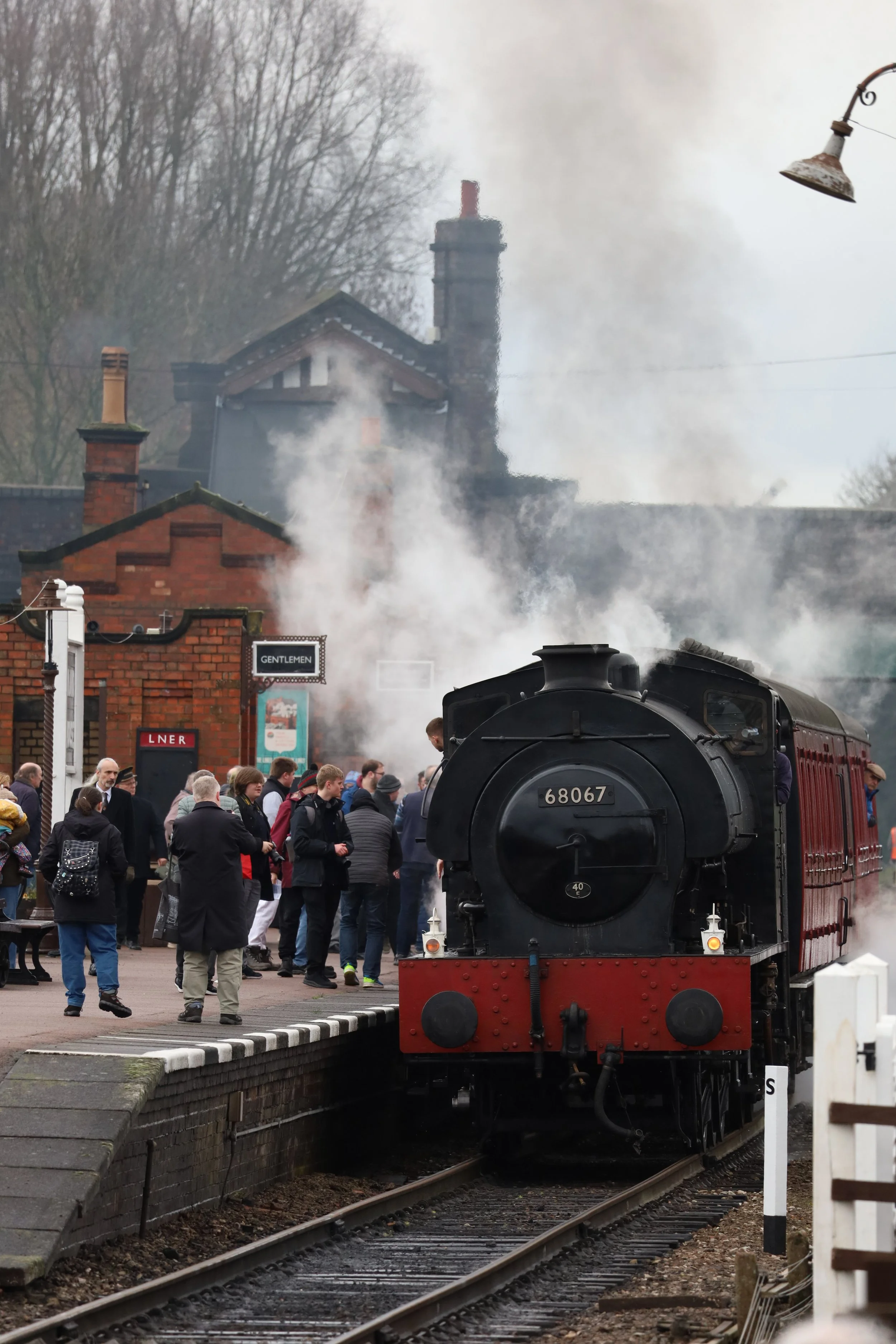 A vintage steam locomotive train on a railway track with a crowd of people gathered nearby, in front of a historic station with brick buildings and leafless trees.
