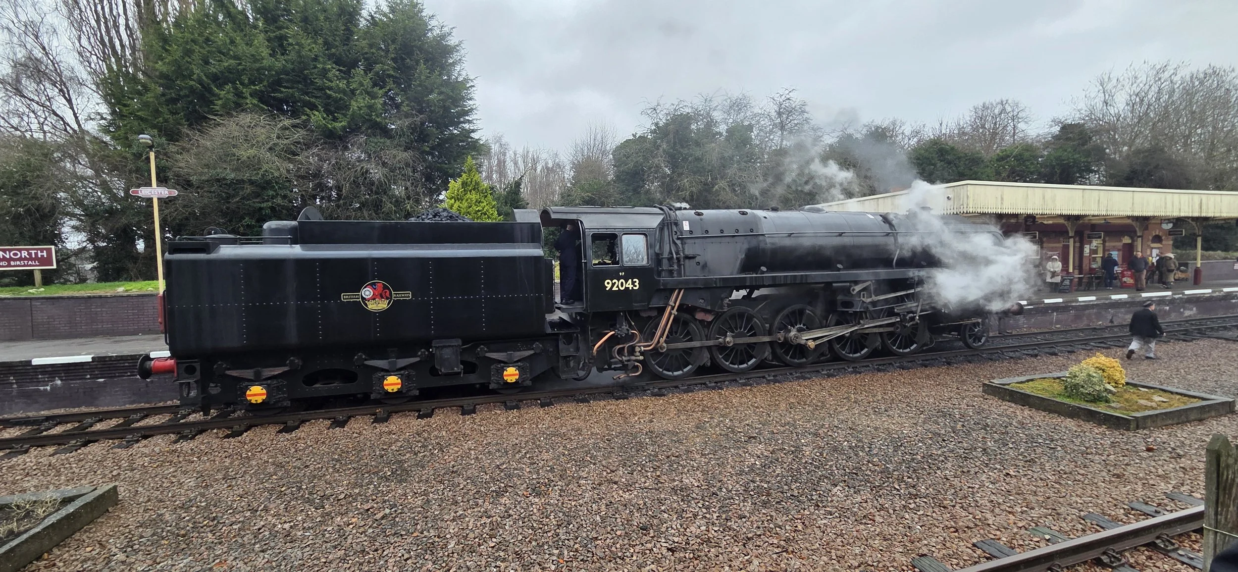 A vintage black steam locomotive on a train track at a station with people waiting on the platform and in the background there are trees and a cloudy sky.