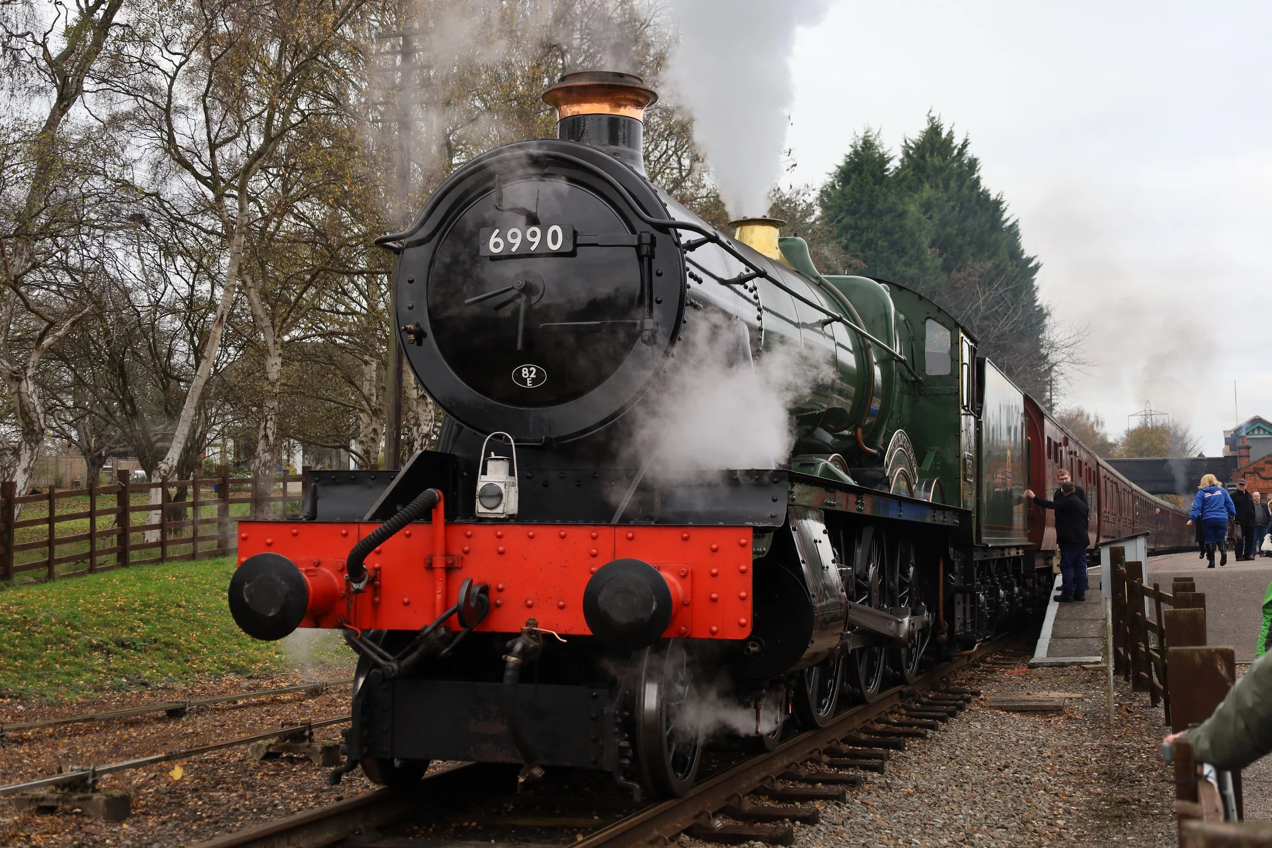 A vintage black steam train with the number 6990 on the front, emitting steam, on railway tracks with people standing nearby, in an outdoor setting with trees and cloudy sky.